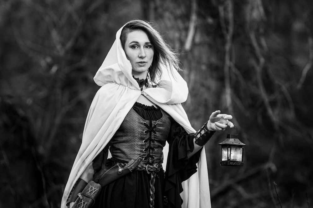 A gothic fantasy-themed black and white portrait of a figure in medieval costume holding a lantern while wearing a hooded cloak.