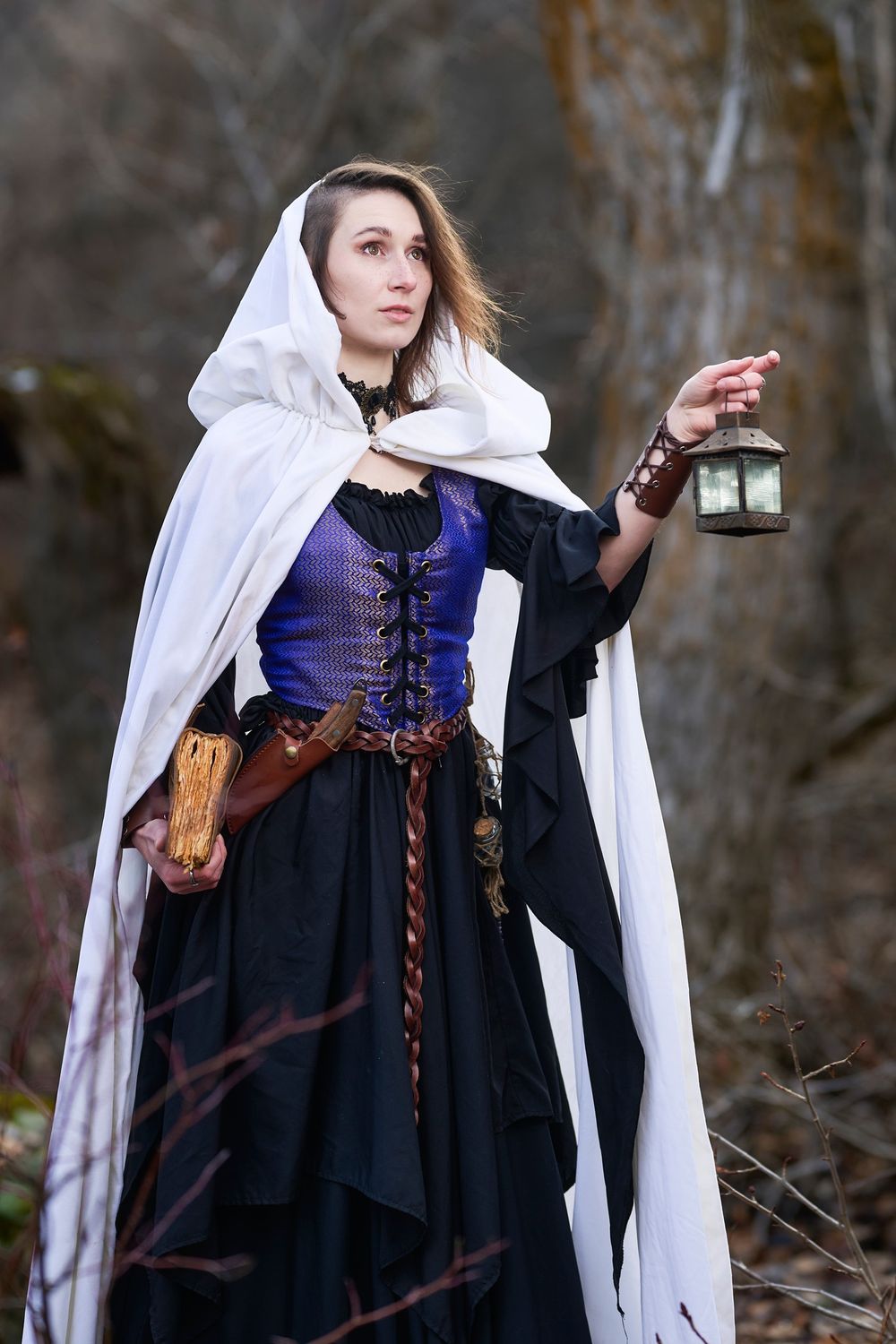 A medieval cosplay featuring a white hooded cloak, corset, and lantern in a moody forest setting.