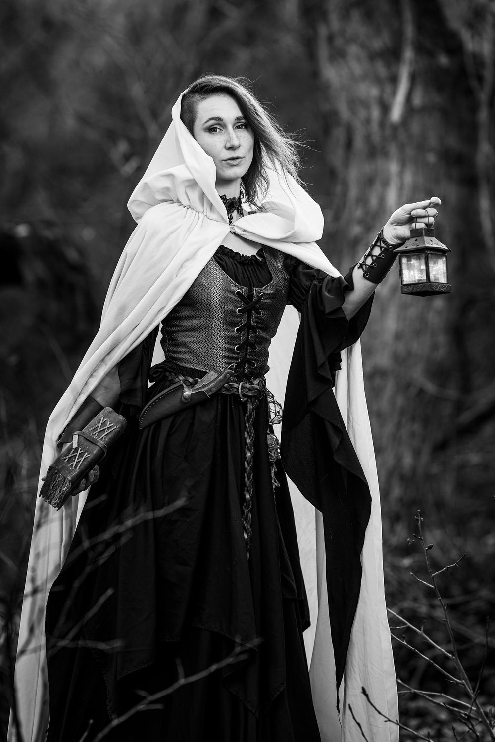 A black and white portrait of someone in medieval attire holding a lantern while wearing a flowing white cloak.