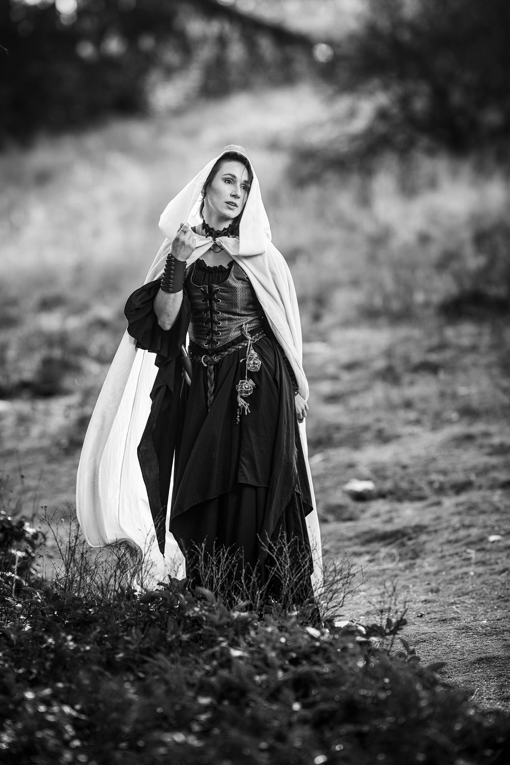 A figure in medieval garb wearing a white hooded cloak and black dress poses on a misty path in black and white.