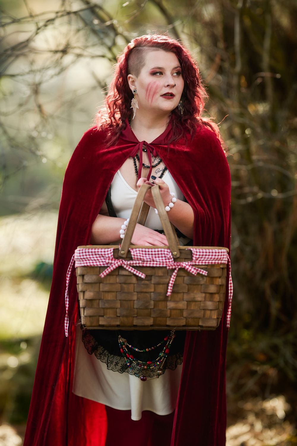 A figure in a bright red hooded cape holds a woven picnic basket with a red and white checkered ribbon in a forest setting.
