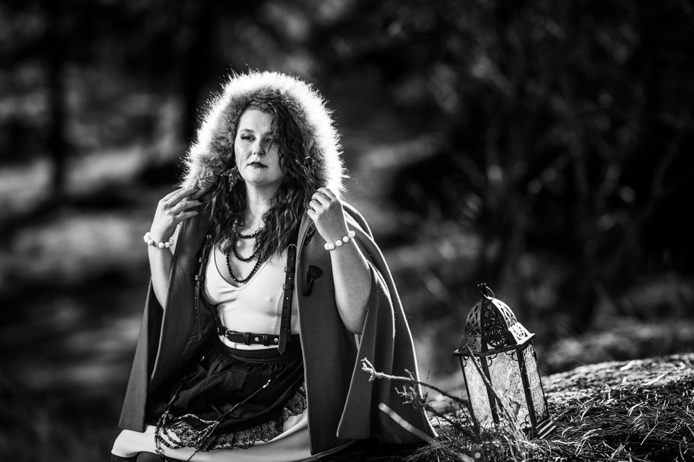 Dramatic black and white portrait of person in fur-trimmed coat sitting outdoors with handbag.