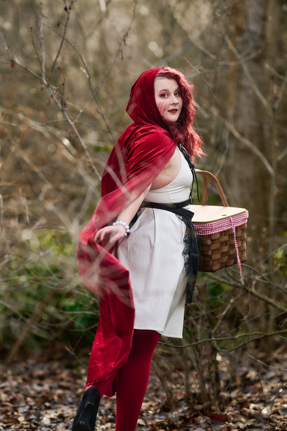 A figure in a red velvet cape and white dress carries a wicker basket through autumn woods.