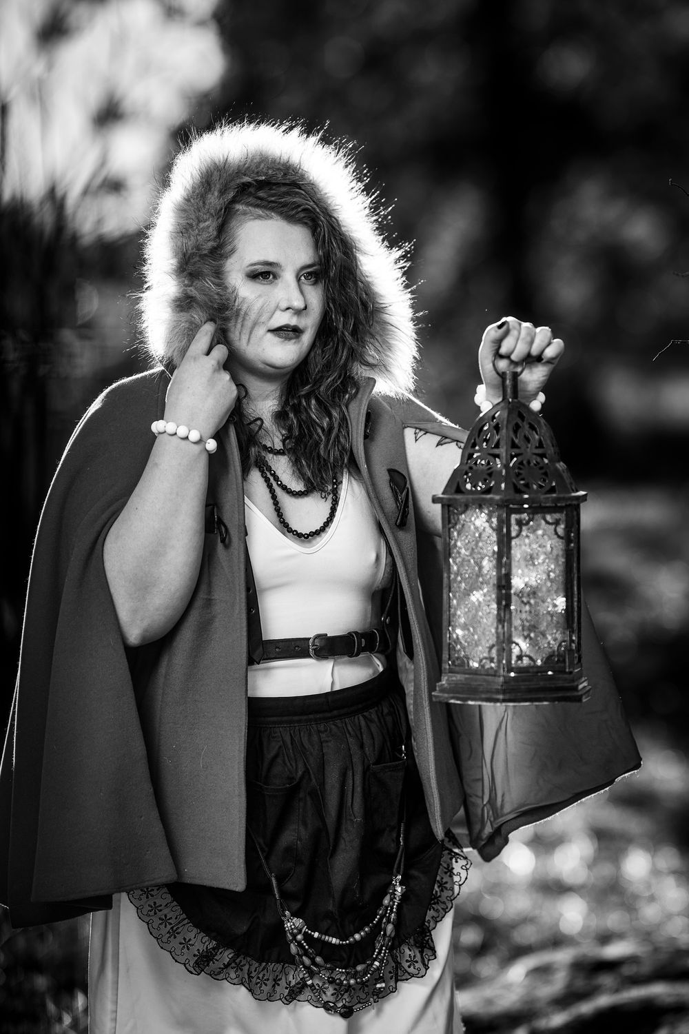 A dreamy black and white portrait of someone holding a decorative lantern while wearing a cape in an ethereal forest setting.