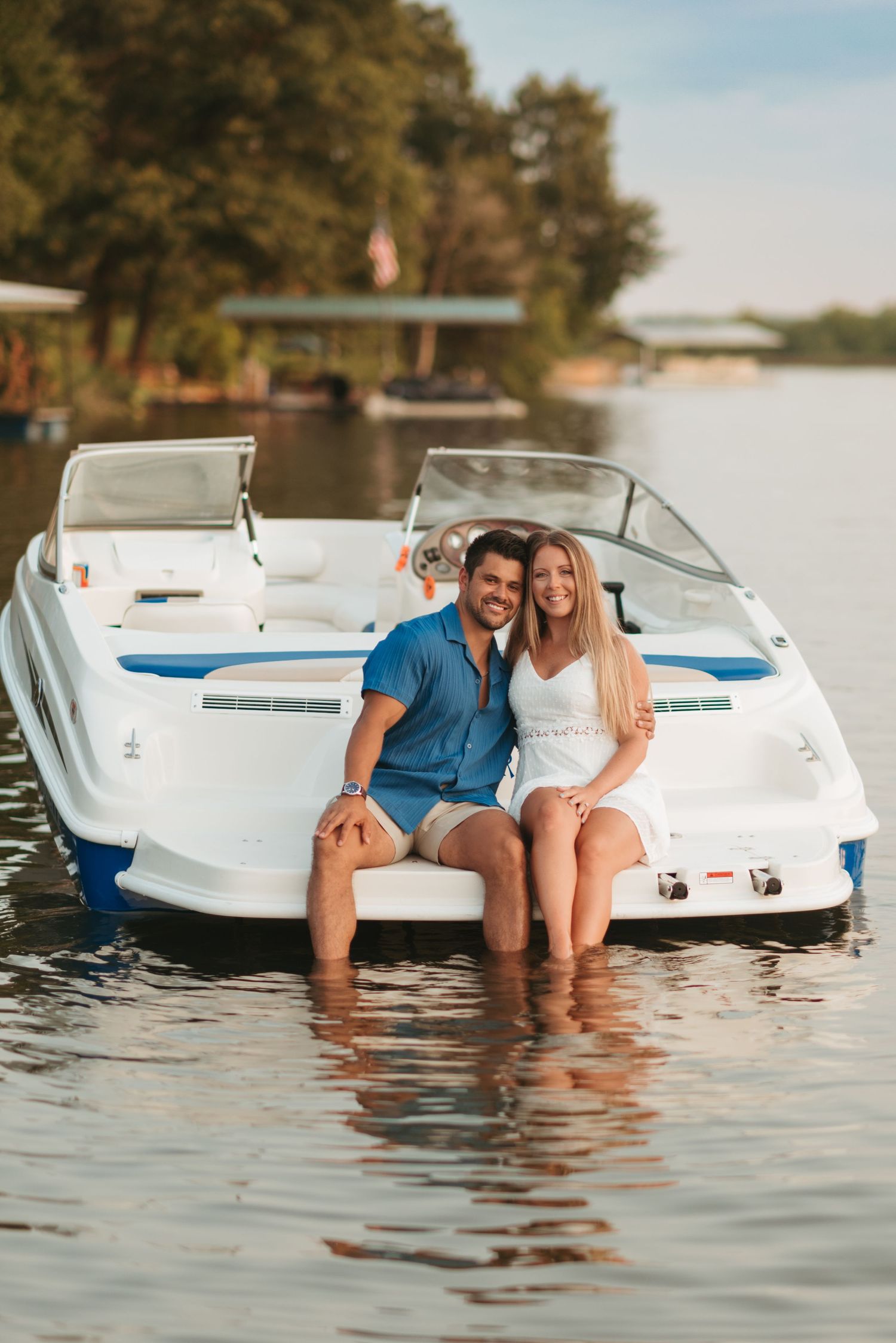 Couple dangling their feet in the water from a vintage white speedboat on a sunny lake.
