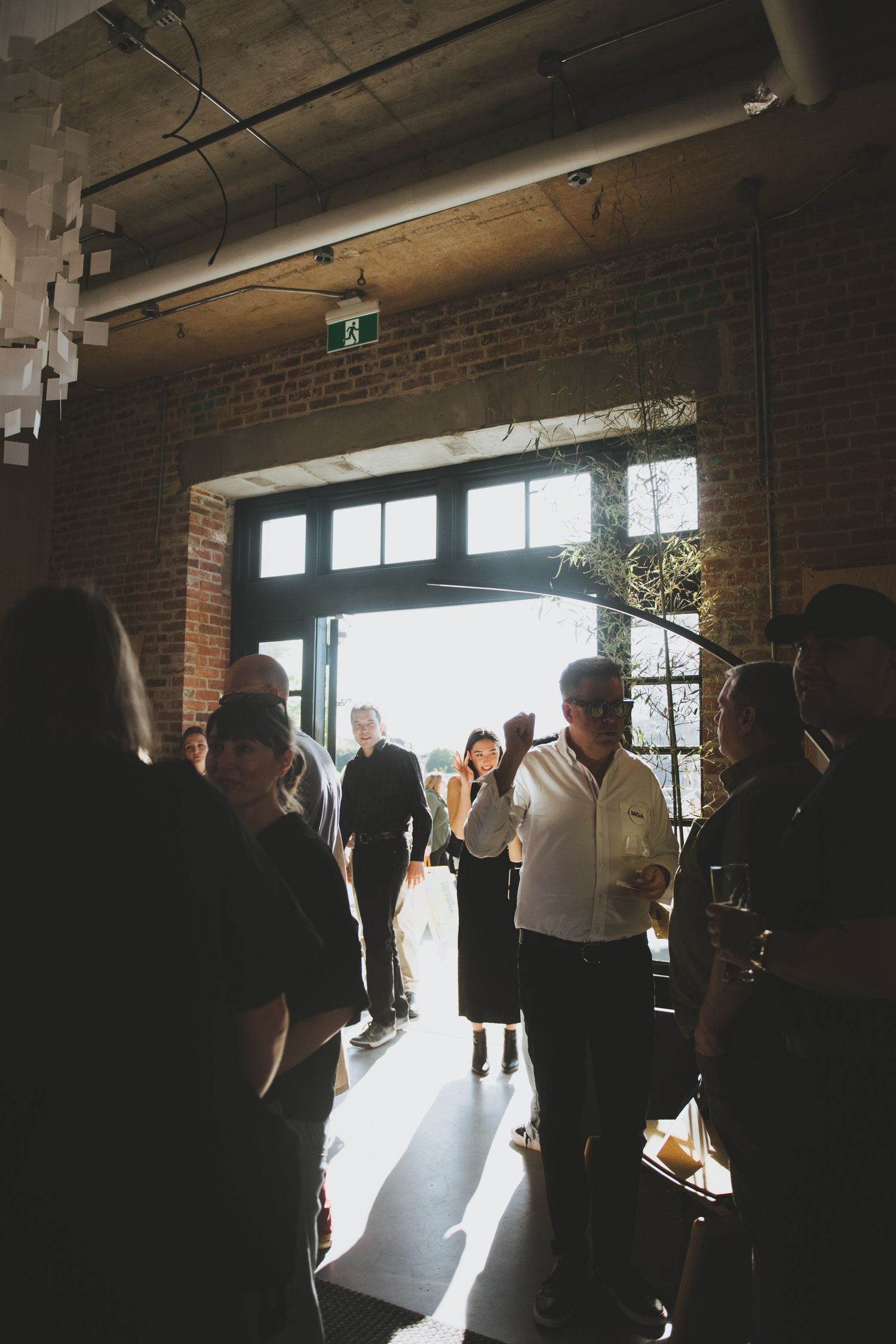 A group of people gather in a sunlit industrial space with exposed brick walls and fluorescent lighting above.