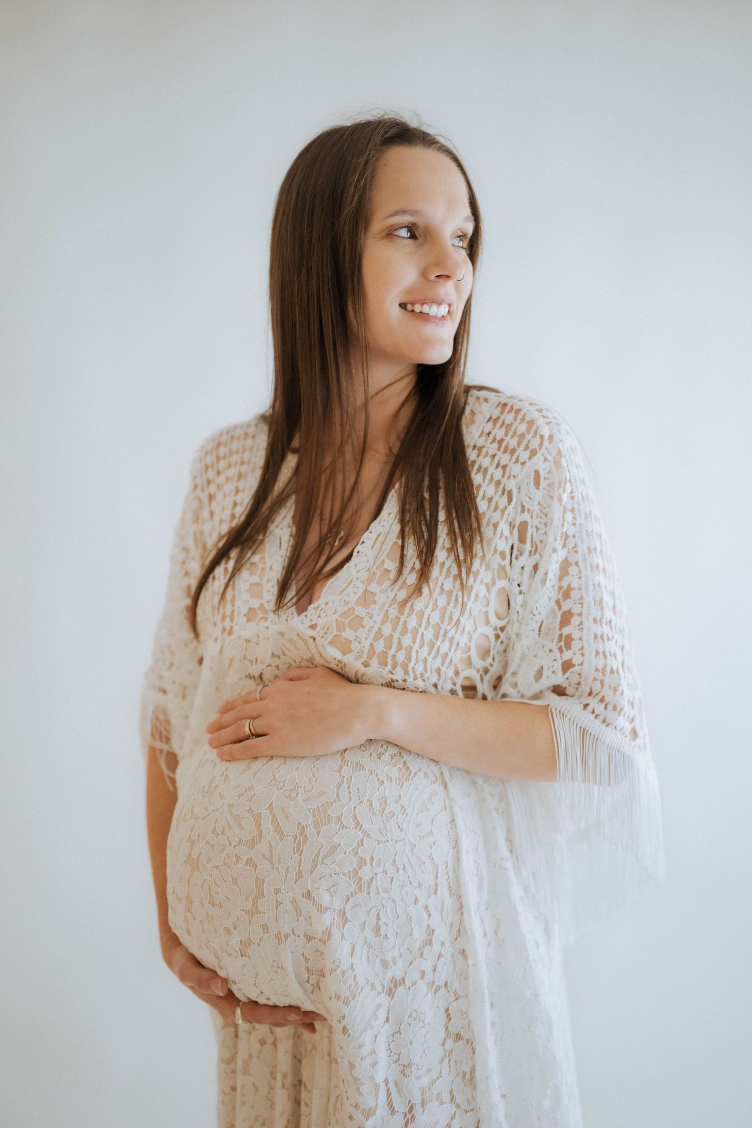 Expectant woman in a white lace maternity dress smiles while standing against light background.