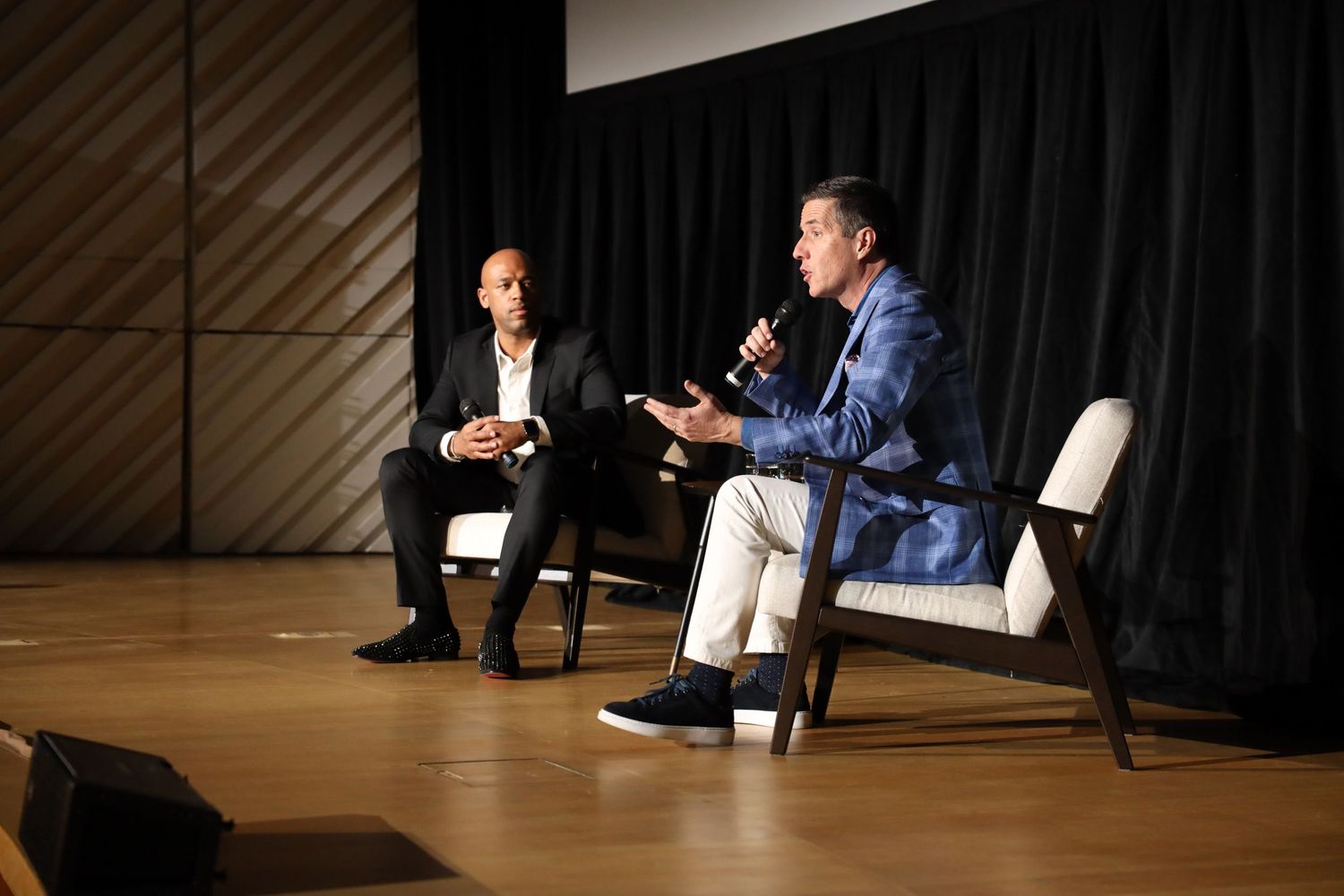 Two people sit in chairs on a stage having a conversation during what appears to be an interview or panel discussion.