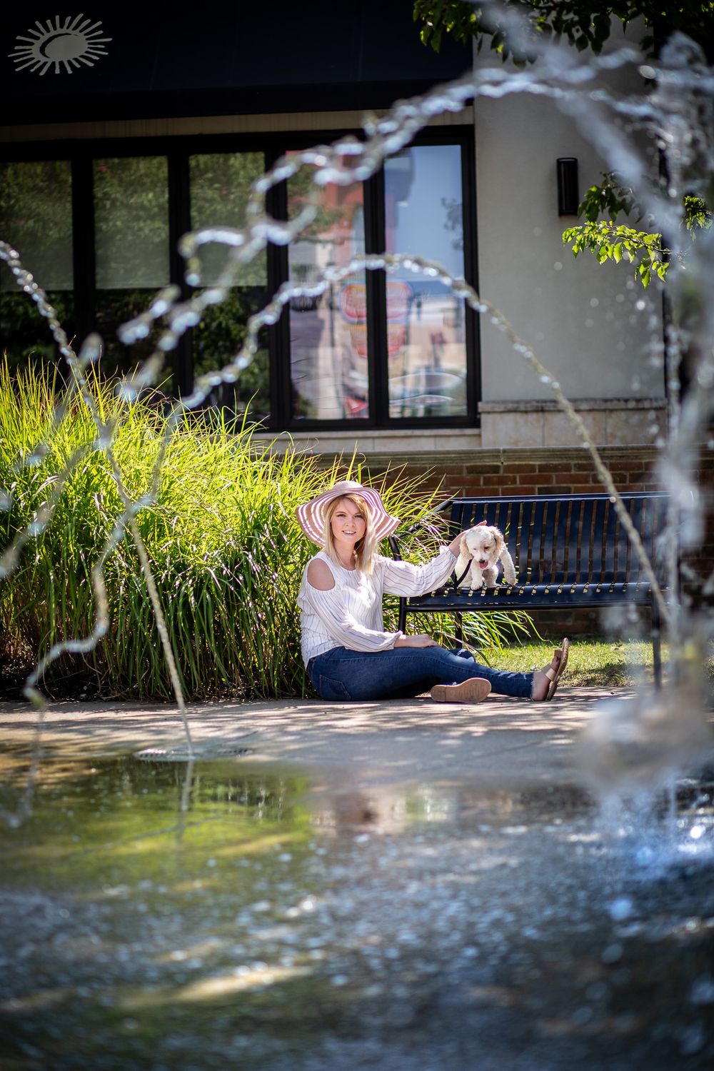 Senior session of a girl with white dog sitting on her lap by a tranquil fountain surrounded by ornamental grasses on a sunny day.