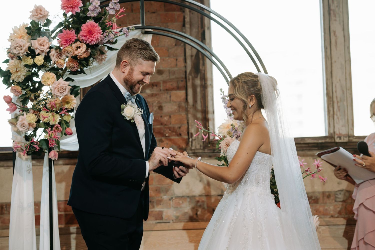 Couple exchanges rings under floral arch during wedding ceremony.