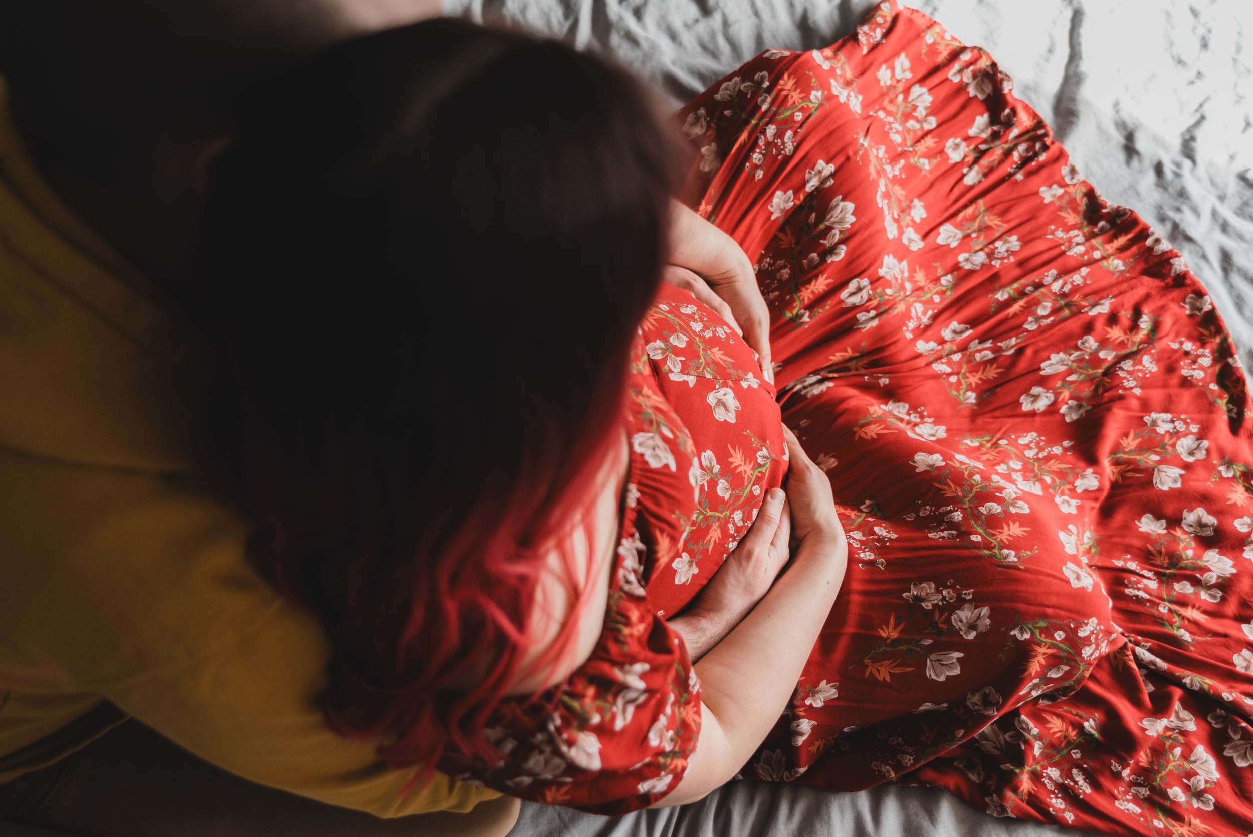 An expectant couple sit on a bed with the mother to be wearing a flowing red floral dress.