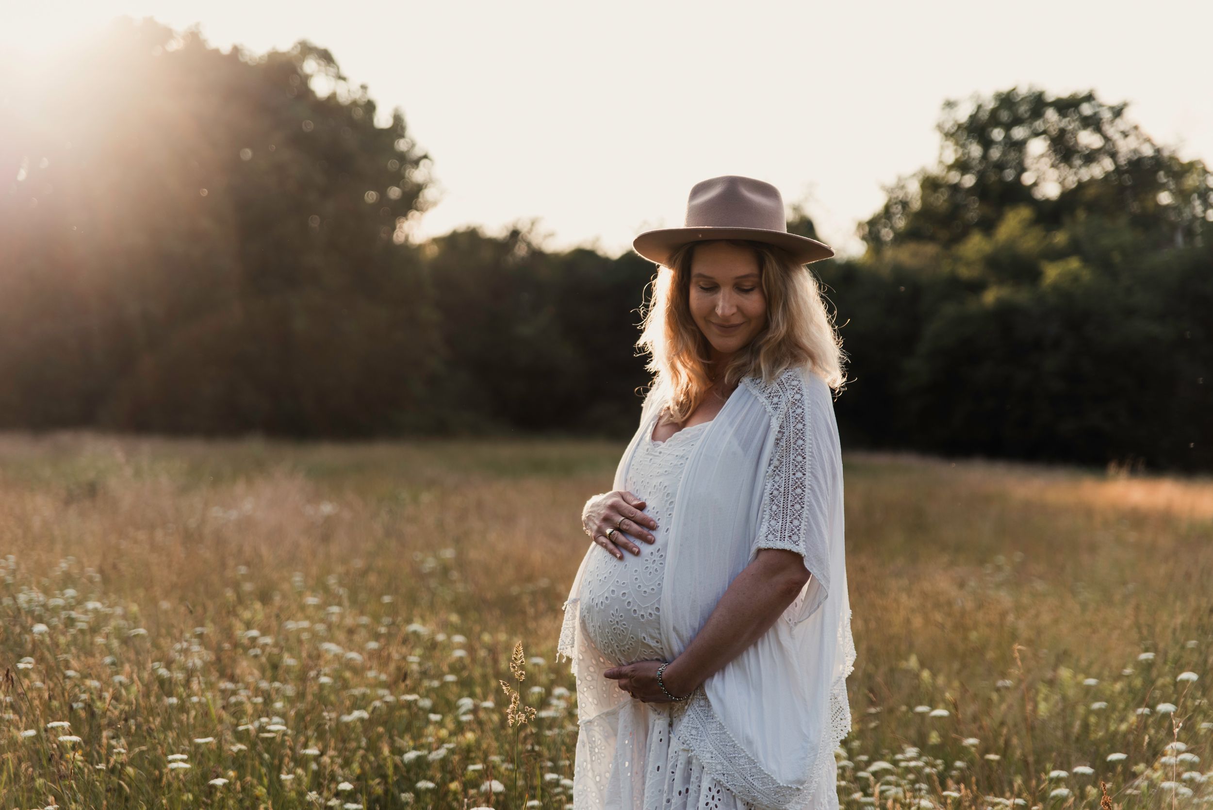 A pregnant woman in a white dress and hat stands in a sunlit wildflower meadow during golden hour.