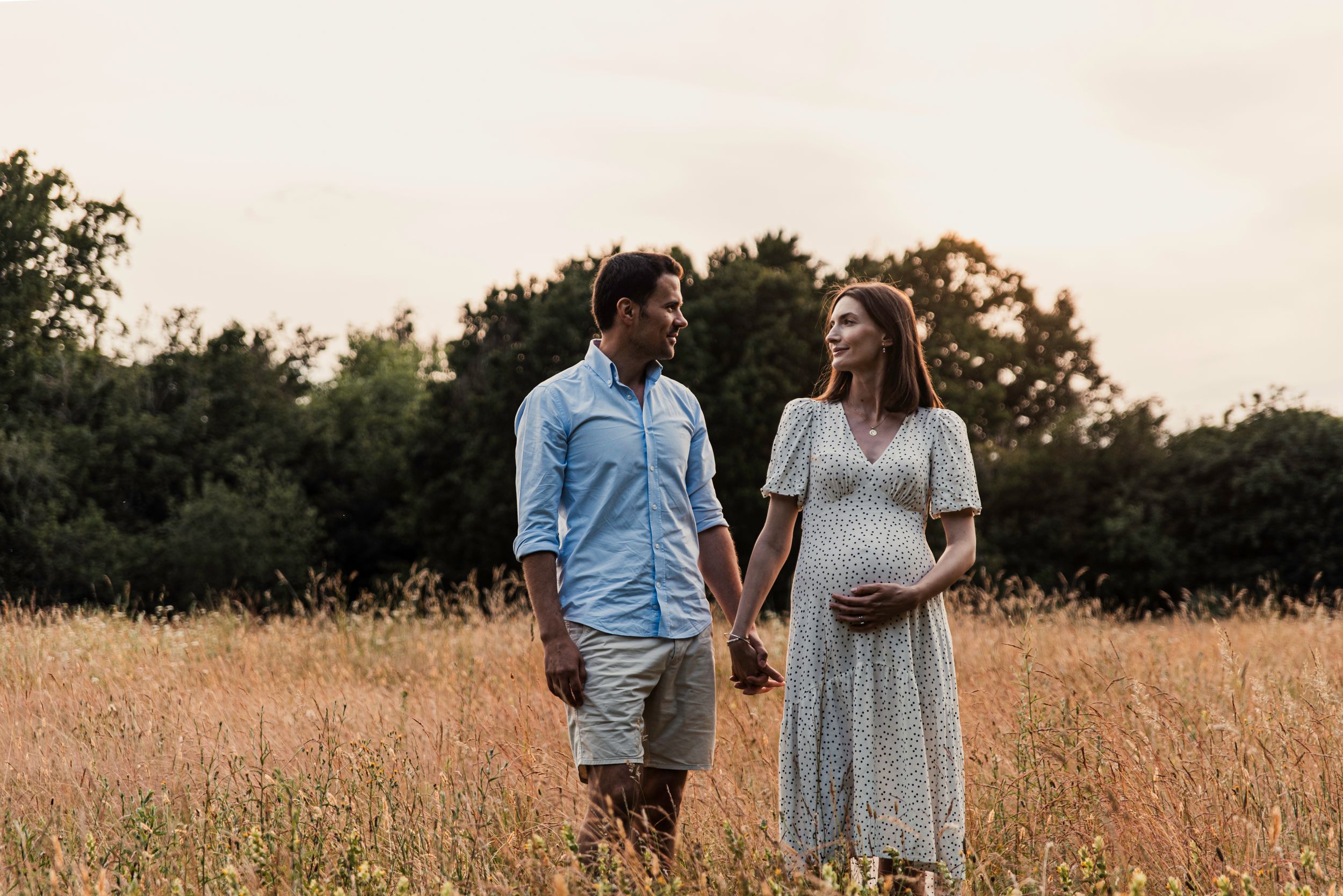 A couple walks hand in hand through a golden field at sunset, with the expectant mother wearing a flowing white dress.