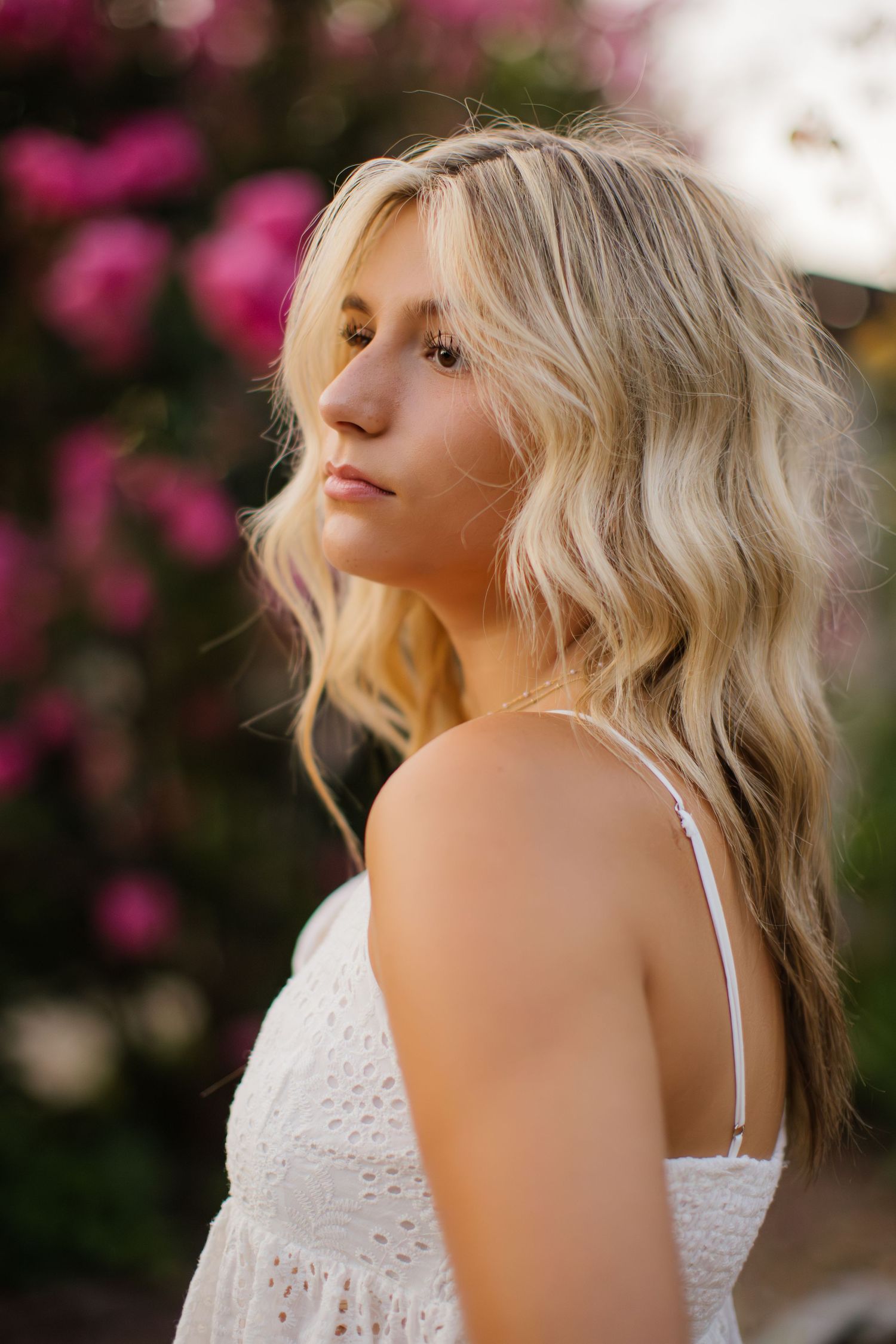 Profile portrait against pink flowers with soft lighting highlighting wavy blonde hair.
