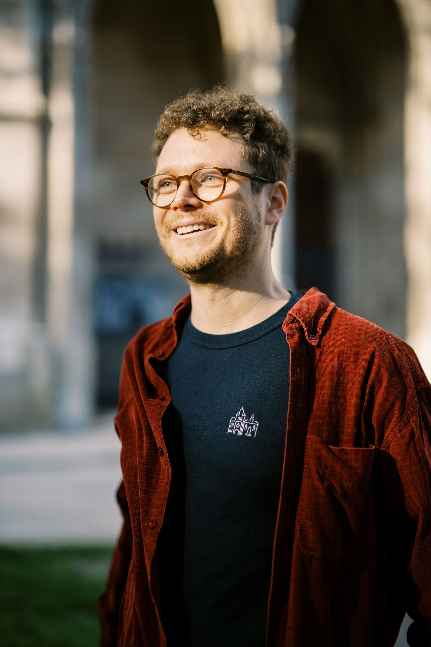 A portrait of someone wearing glasses and a burgundy corduroy jacket, smiling warmly in natural outdoor lighting.