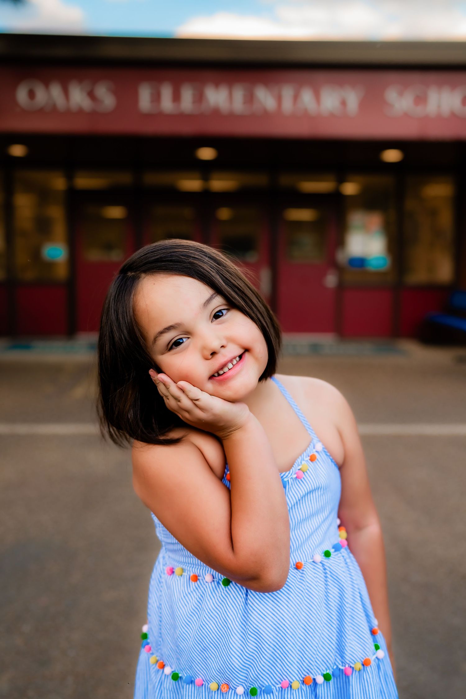 Child in blue summer dress posing playfully outside elementary school entrance.
