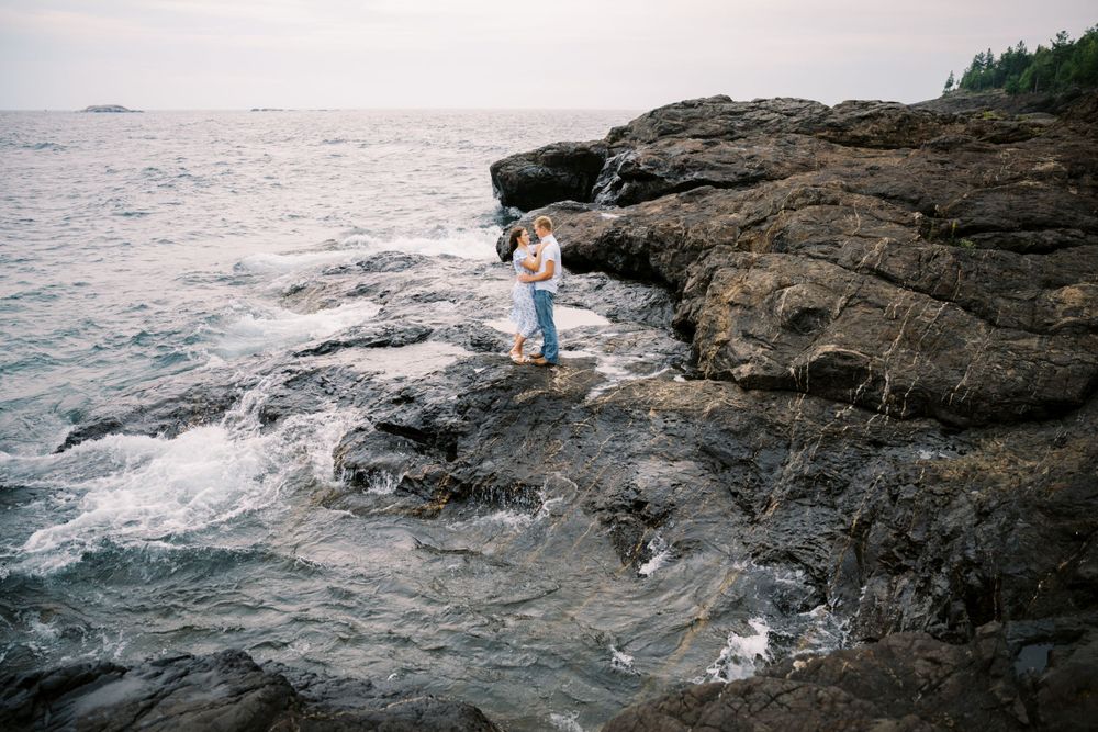 A figure stands on rugged coastal rocks as ocean waves crash against the rocky shoreline at sunset.
