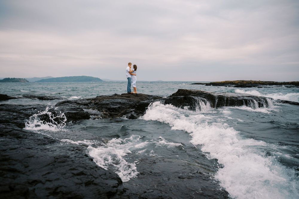 A couple embraces on dramatic rocky coastline as ocean waves crash against the shore at dusk.