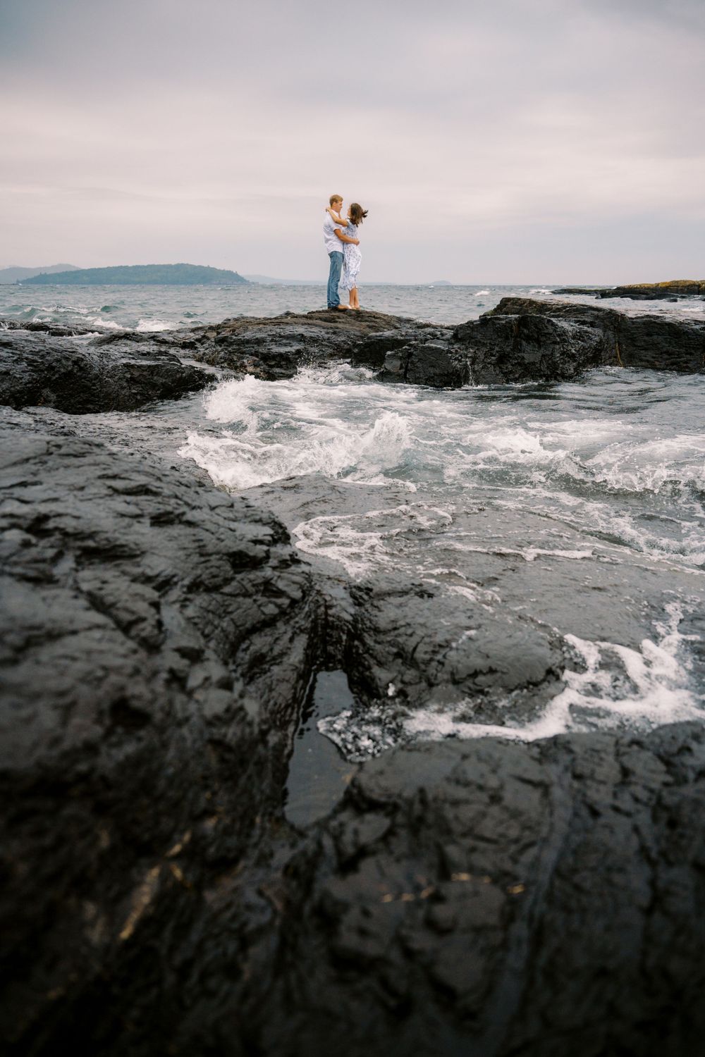 Two figures stand embracing on dramatic rocky coastline as waves crash against dark cliffs at sunset.