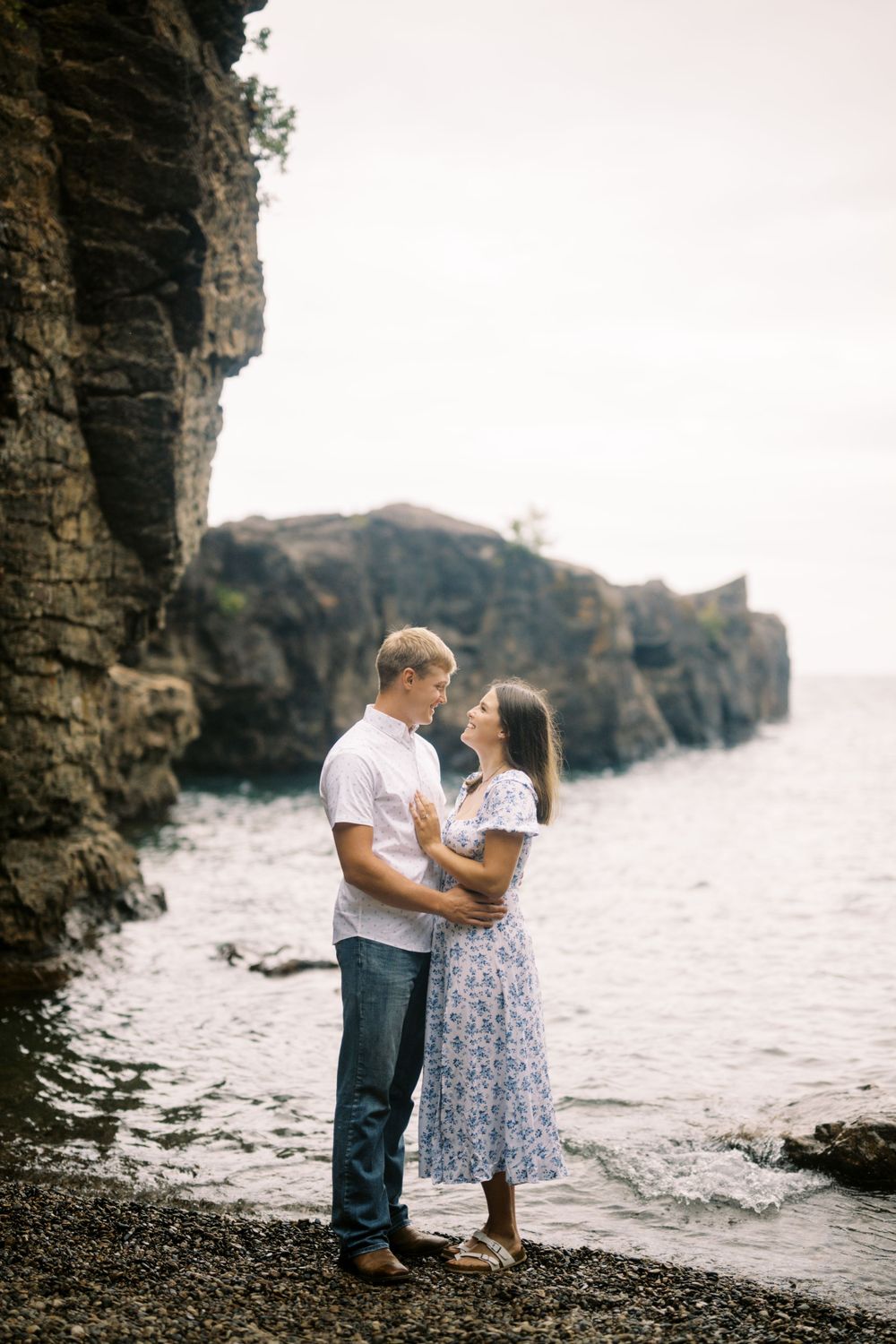 A couple embraces on a rocky beach with dramatic cliffs and crashing waves in the background.