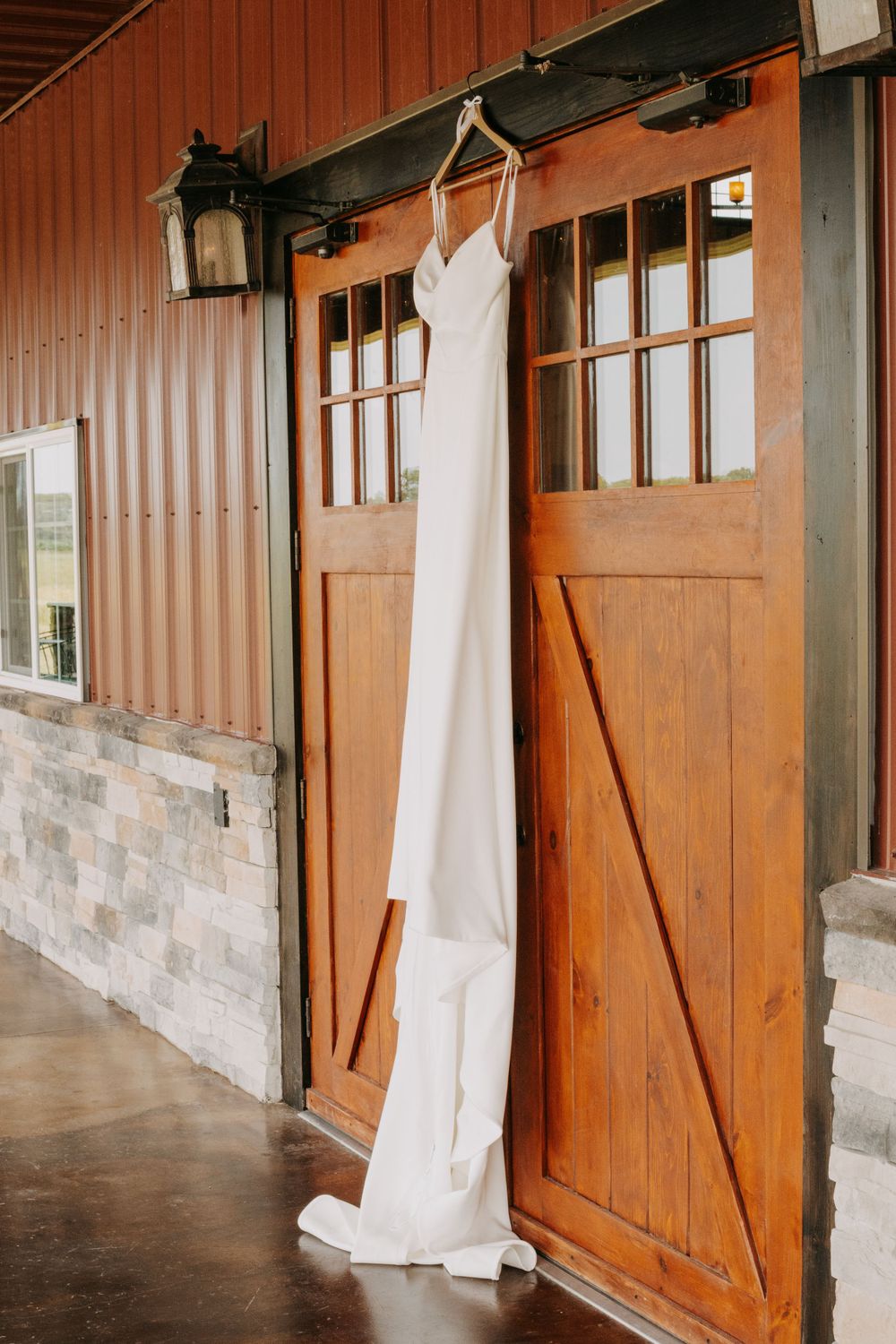 Elegant white wedding dress hanging on wooden barn door with lantern fixture above.