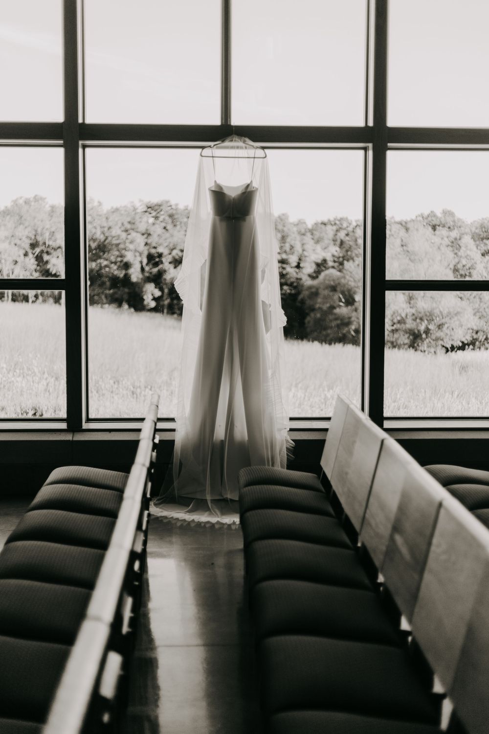 A white wedding dress hangs in front of large windows with empty black chairs in a minimalist black and white composition.