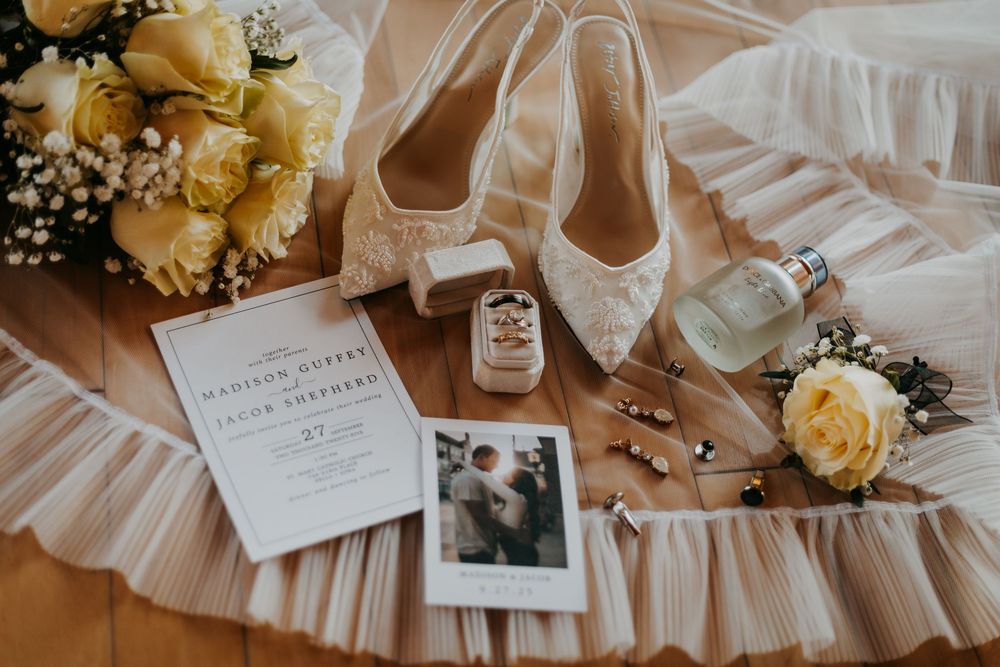 Wedding details featuring delicate cream roses, invitation cards, and glassware arranged on a soft white fabric backdrop.