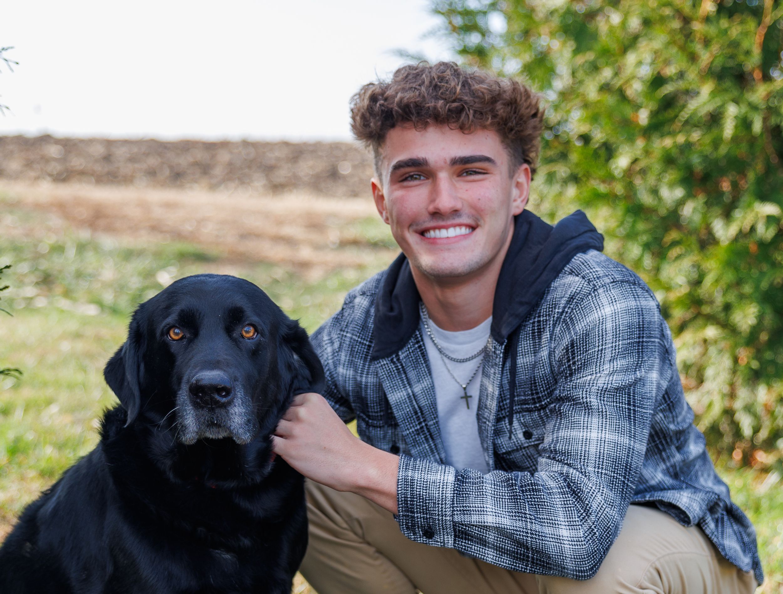 A black Labrador retriever poses with someone in a plaid shirt outdoors against a green leafy background.