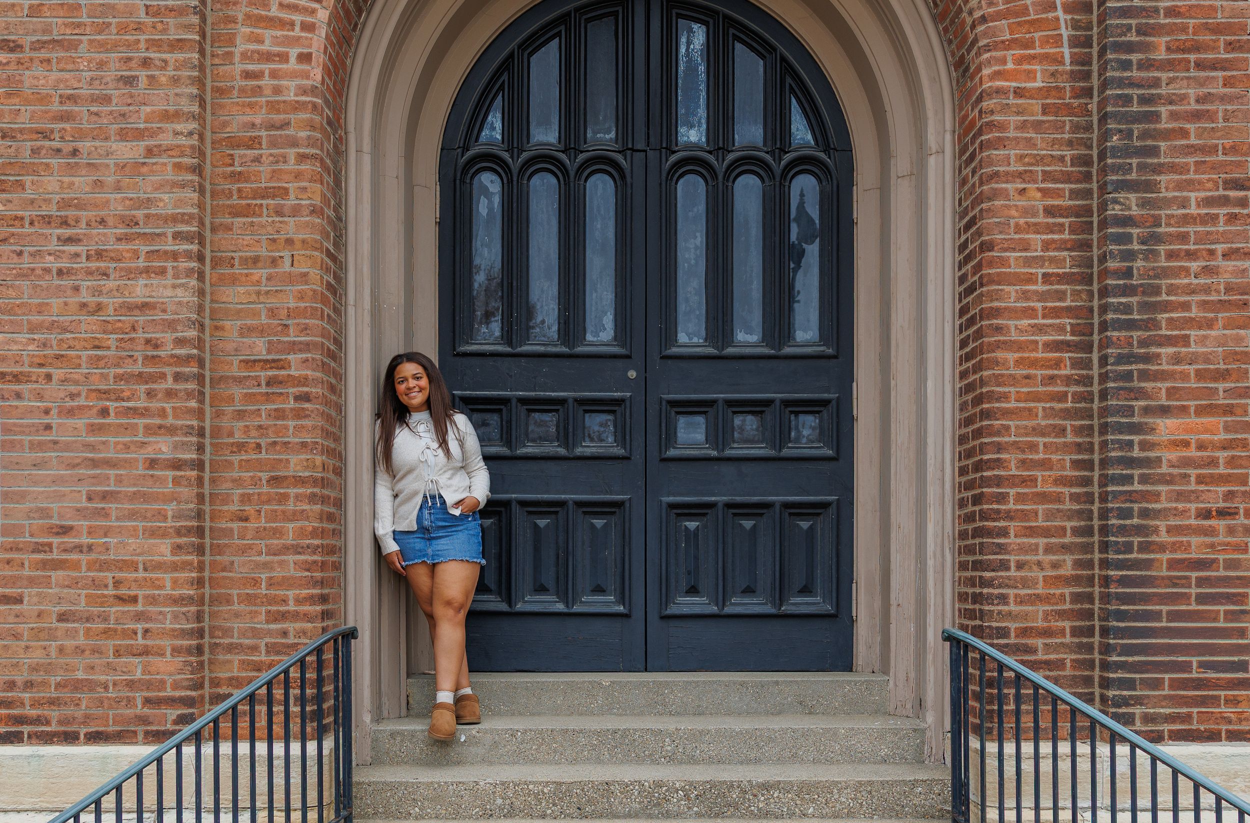 Someone poses in a white top and blue shorts at the entrance of a brick building with an arched doorway.