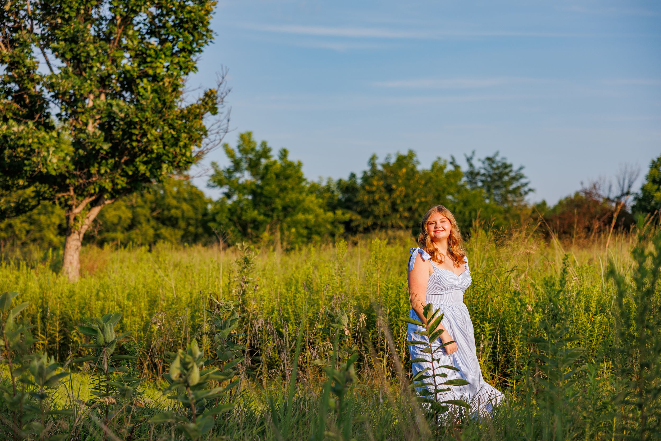 A person in a white dress sits in a golden meadow during sunset while surrounded by tall summer grasses.