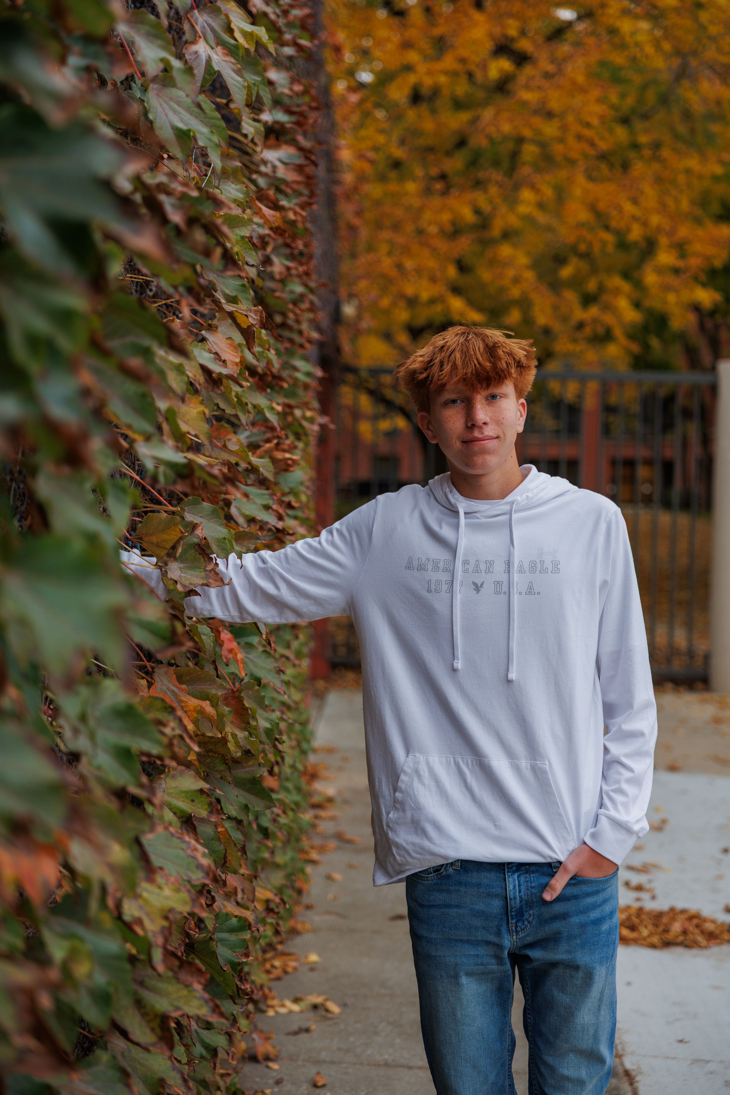 A person in a white hoodie and blue jeans stands outdoors against a backdrop of autumn trees with orange foliage.