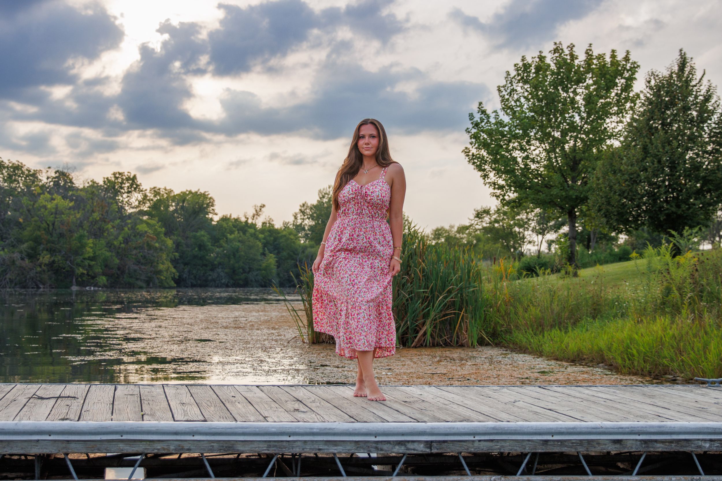 A person in a pink dress stands on a wooden dock at sunset near a peaceful lake.