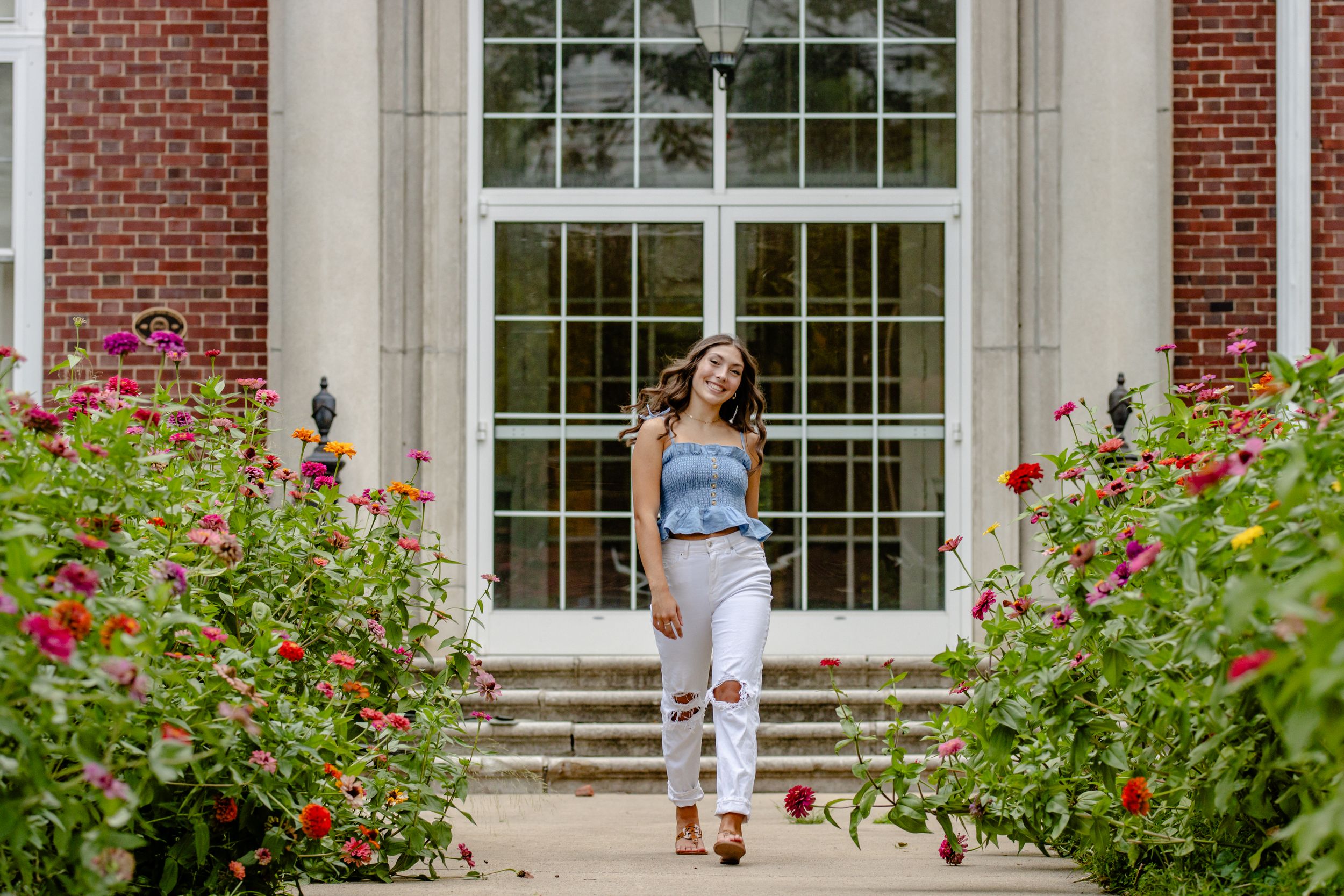 Someone in casual attire poses in front of a building with red flowers blooming.