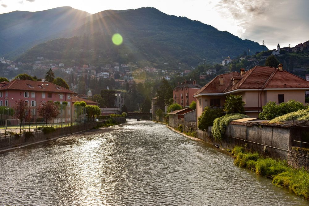 Golden sunset rays burst over mountain town with river canal lined by colorful European buildings in Lugano, Switzerland.