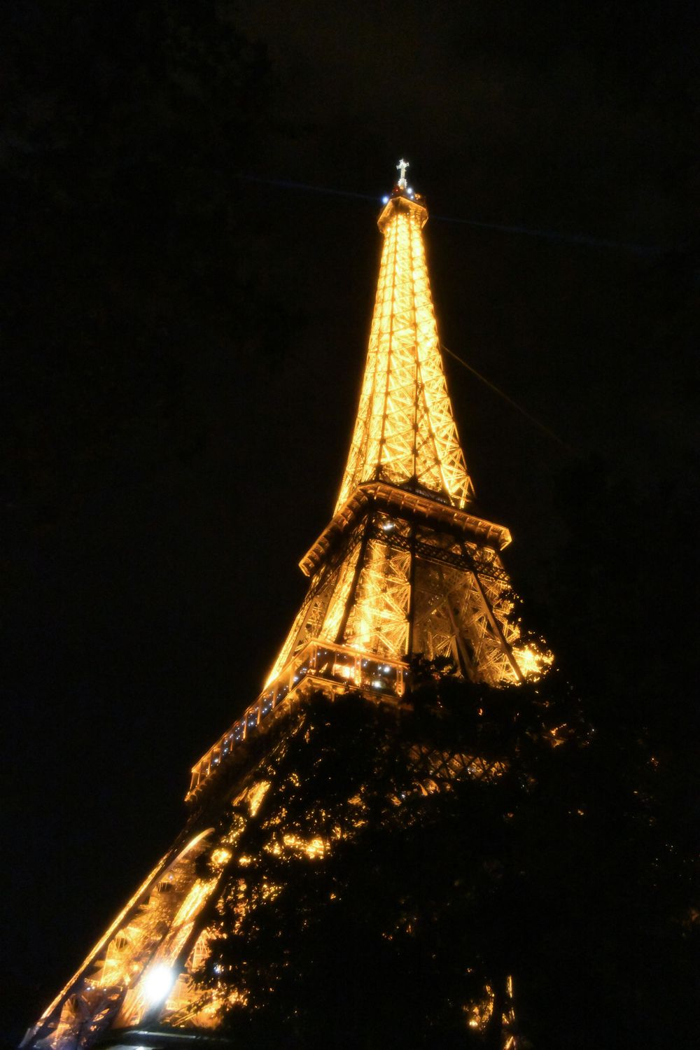 Low-angle view of the illuminated Eiffel Tower at night with golden lights shining through surrounding trees.