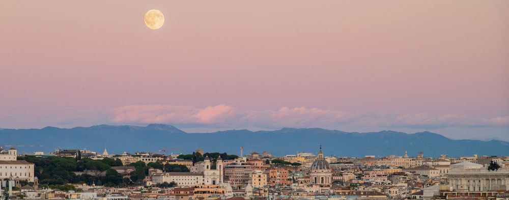 Full moon rising over Rome skyline at dusk with pink sky and mountain backdrop.