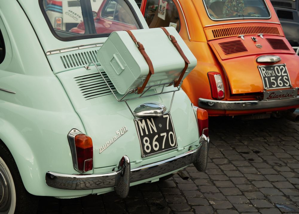 Two vintage Fiat 500 cars, mint green with luggage rack and orange, parked on cobblestones in Italy.