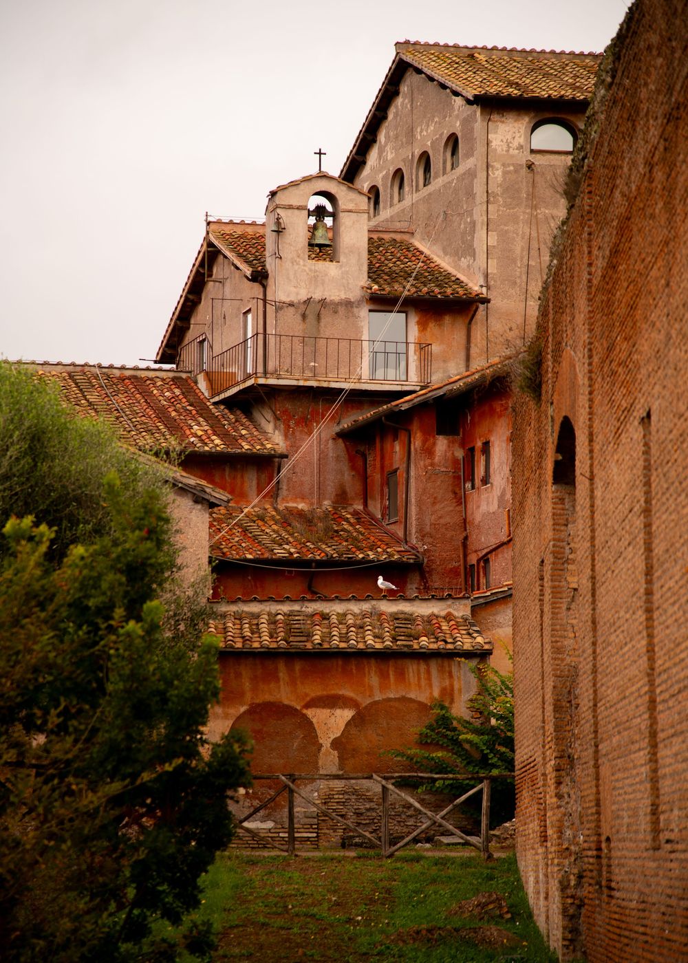 Ancient Roman brick buildings with terracotta rooftops, arched windows, and a small bell tower under an overcast sky.