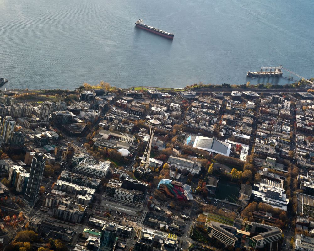 Aerial view of a dense urban cityscape near a waterfront with ships anchored in calm blue waters.