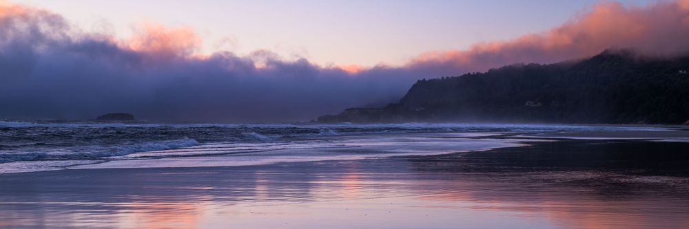 Dramatic sunset over a misty coastal beach with pink clouds reflecting on wet sand and dark forested hills in the background.