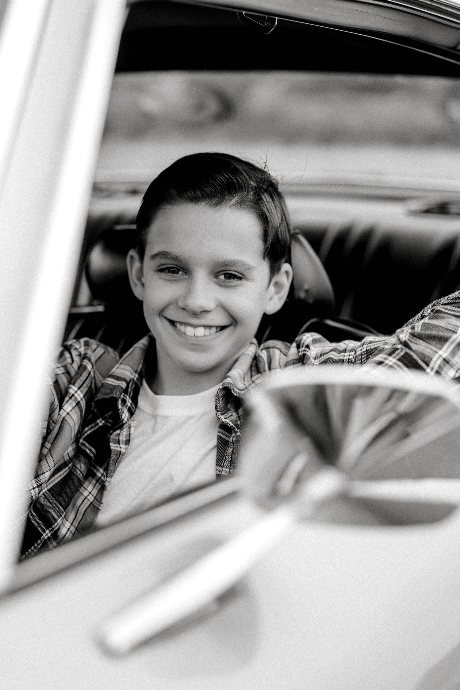 Smiling young boy sits inside a classic vintage car, wearing a plaid shirt, black and white photo.