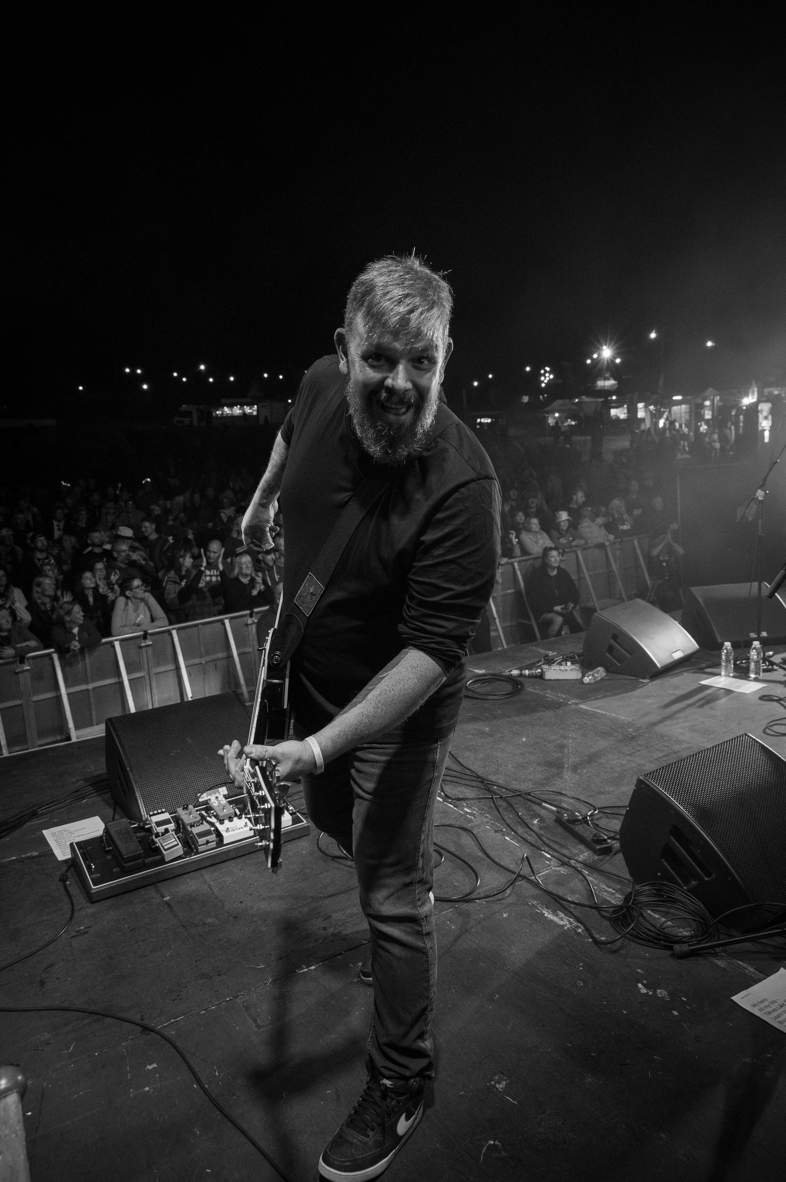 Musician adjusts pedal board on dark stage with city lights visible in background.