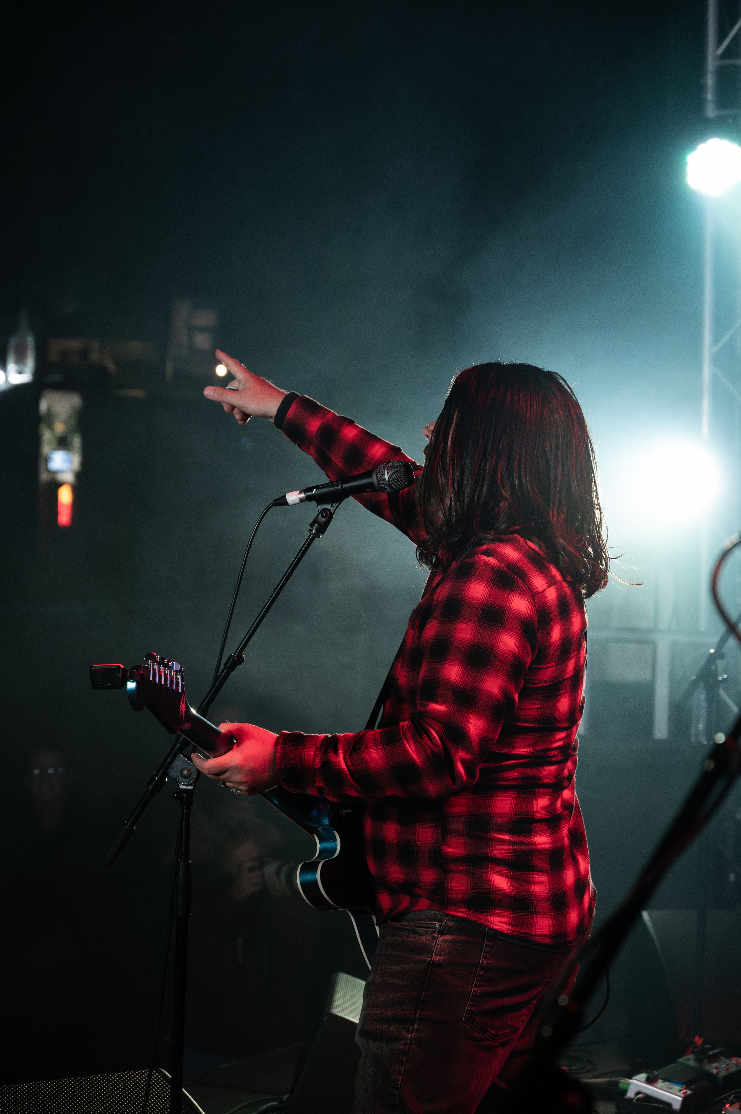 Performer in red plaid flannel shirt points while singing on dimly lit stage.