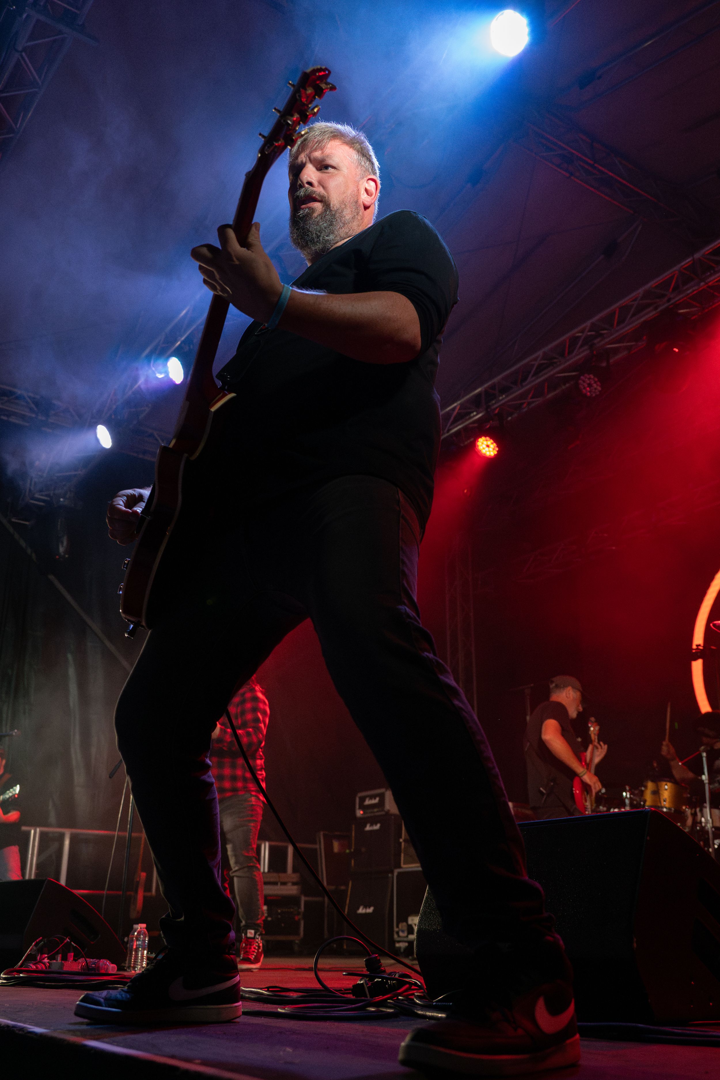 Guitarist in black performs energetically on stage with dramatic red and blue lighting.