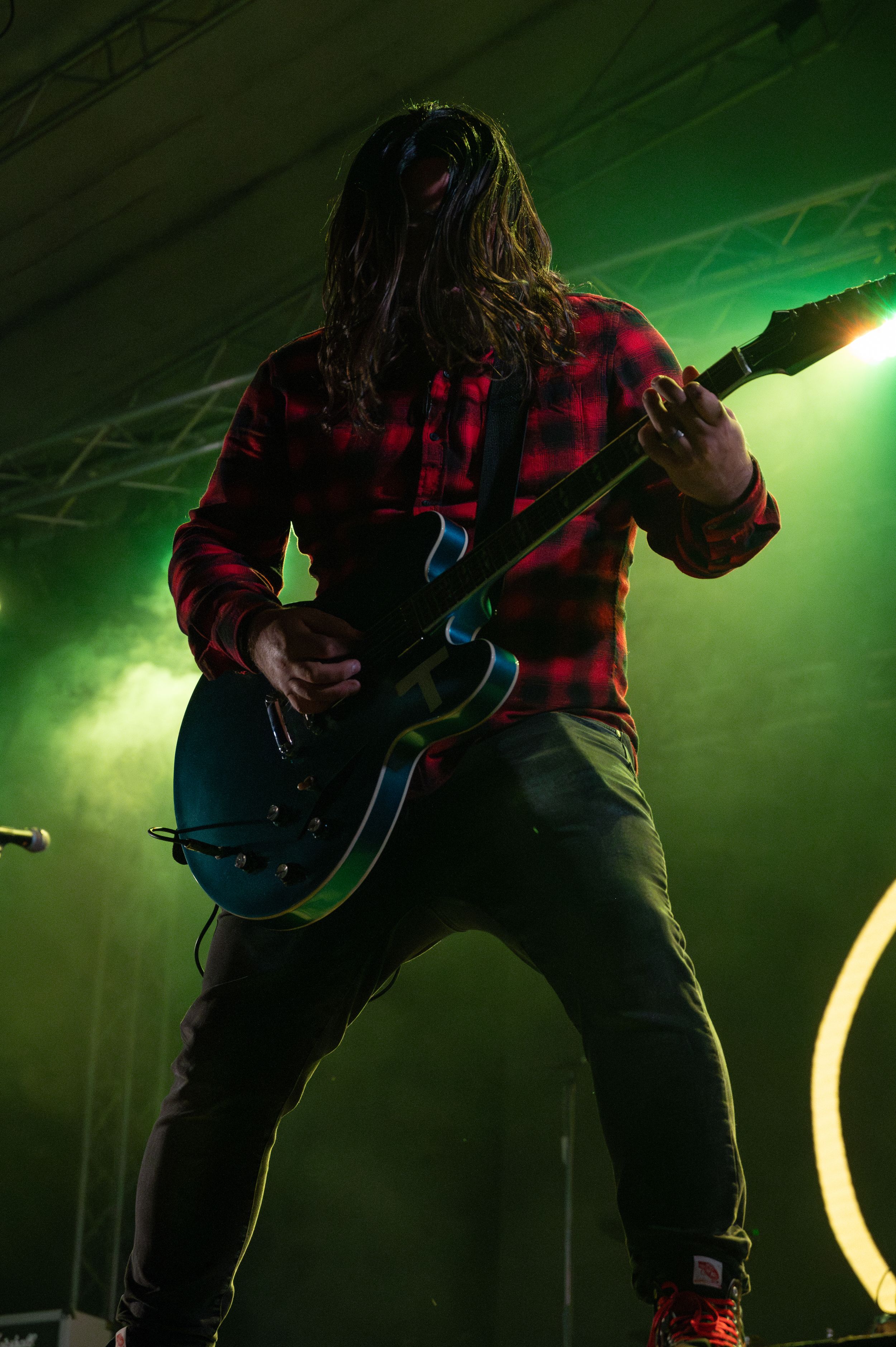 Guitarist in red plaid shirt silhouetted against green stage lighting during performance.