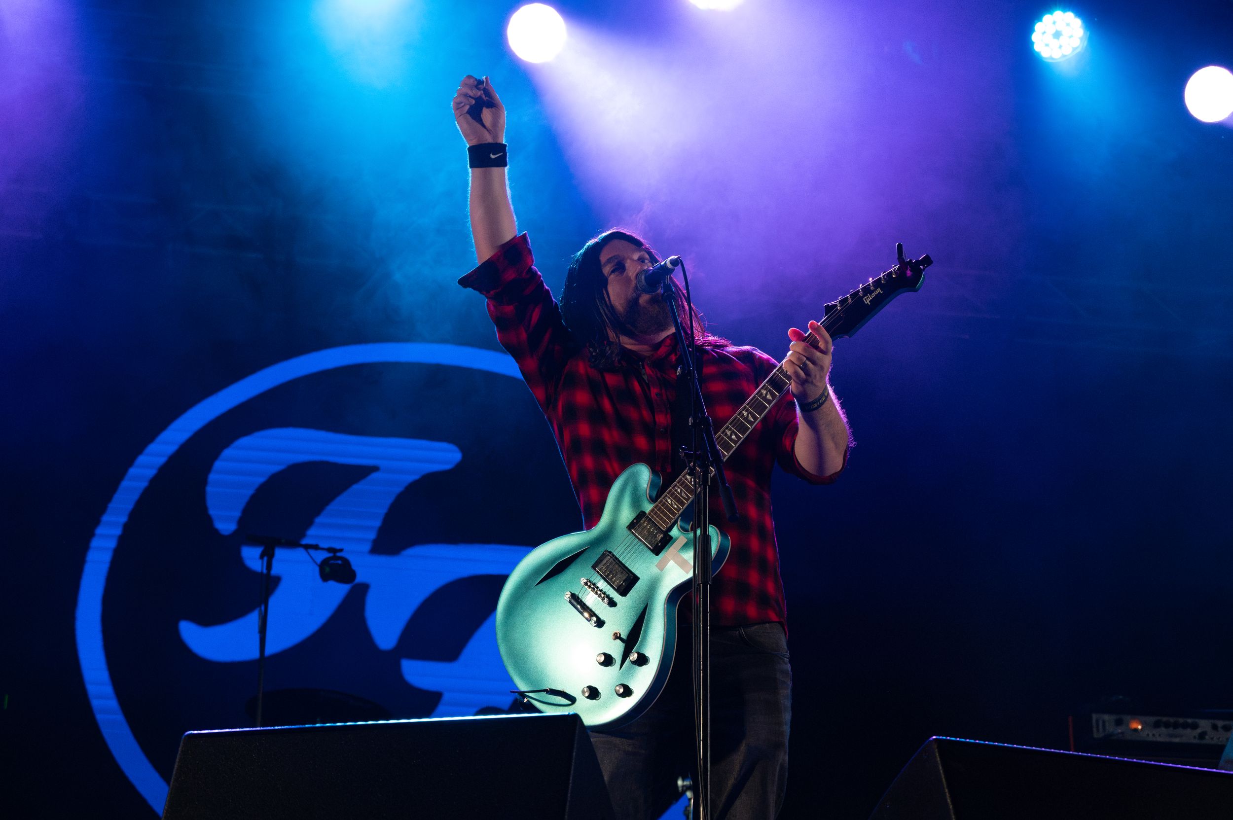 Guitarist performs under blue stage lights with band logo visible behind.