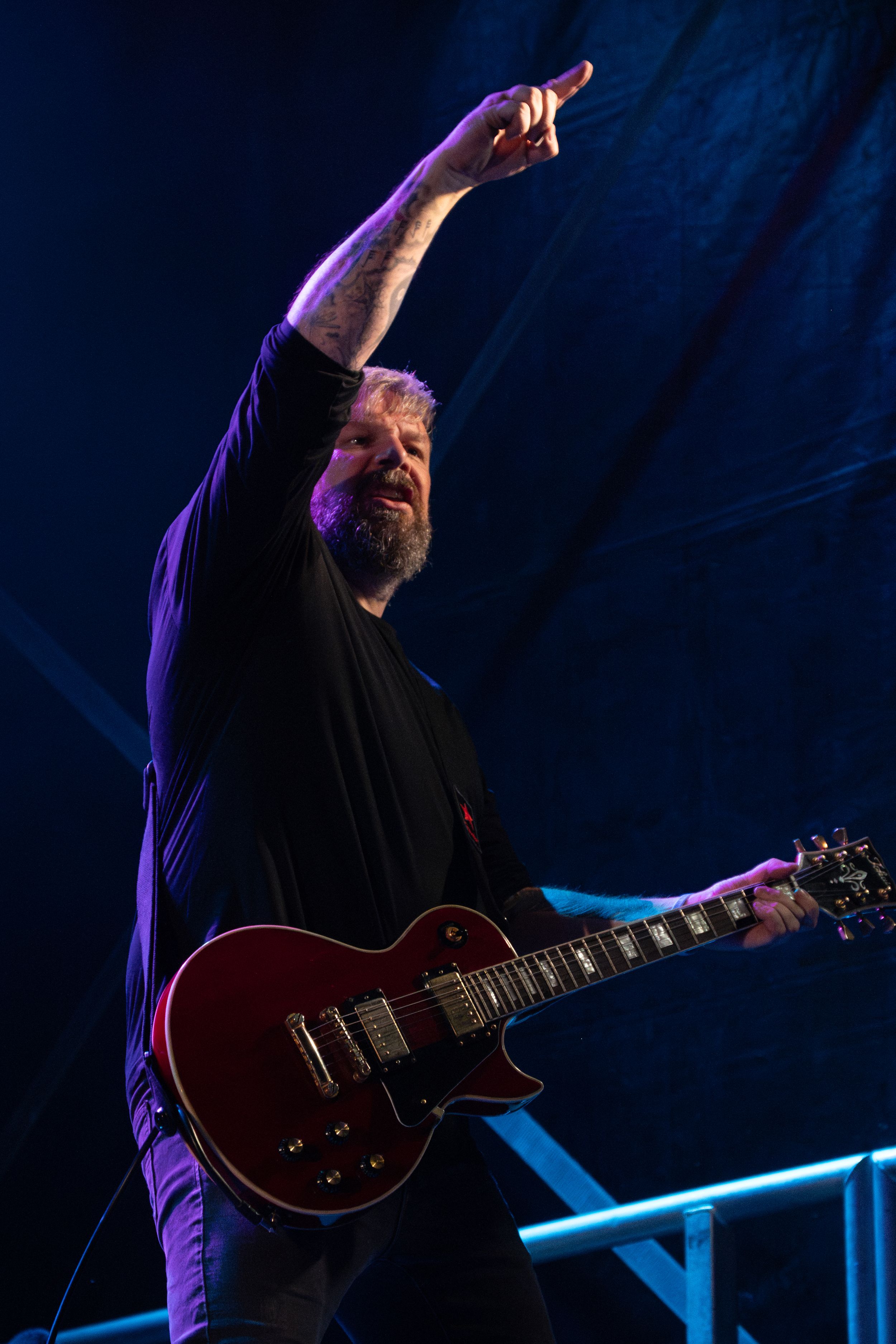 Musician performs on stage with a red electric guitar while pointing upward under dramatic purple lighting.