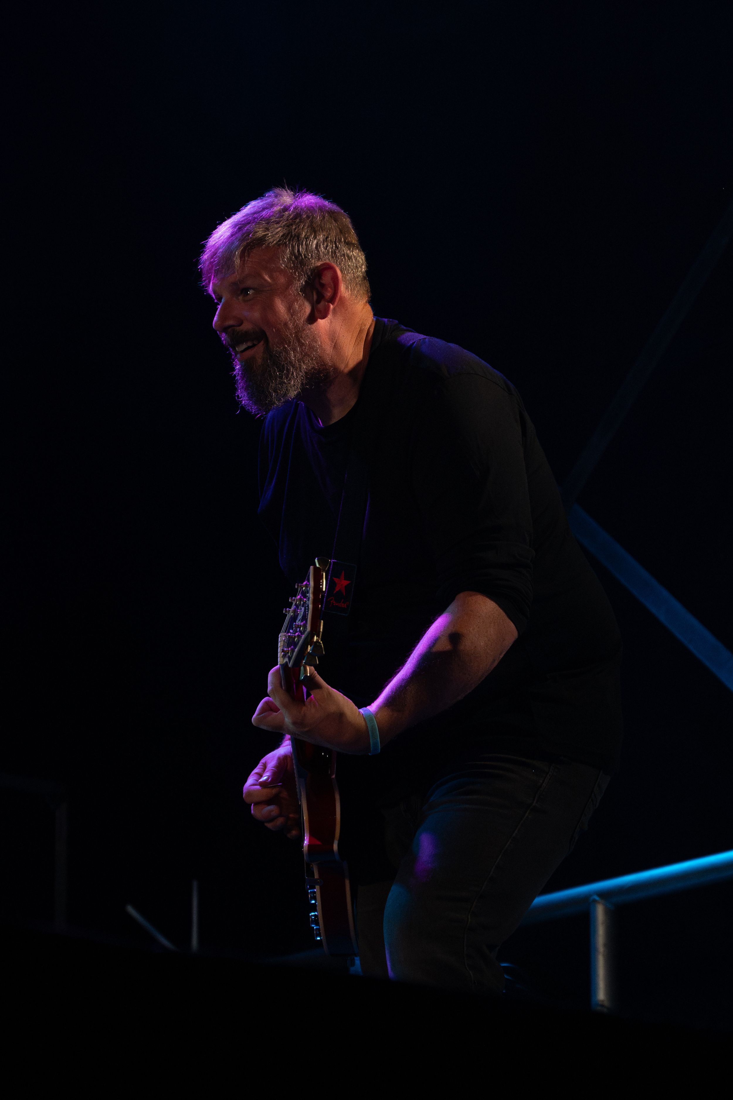 Guitarist in black clothing plays on darkened stage illuminated by purple stage lights.