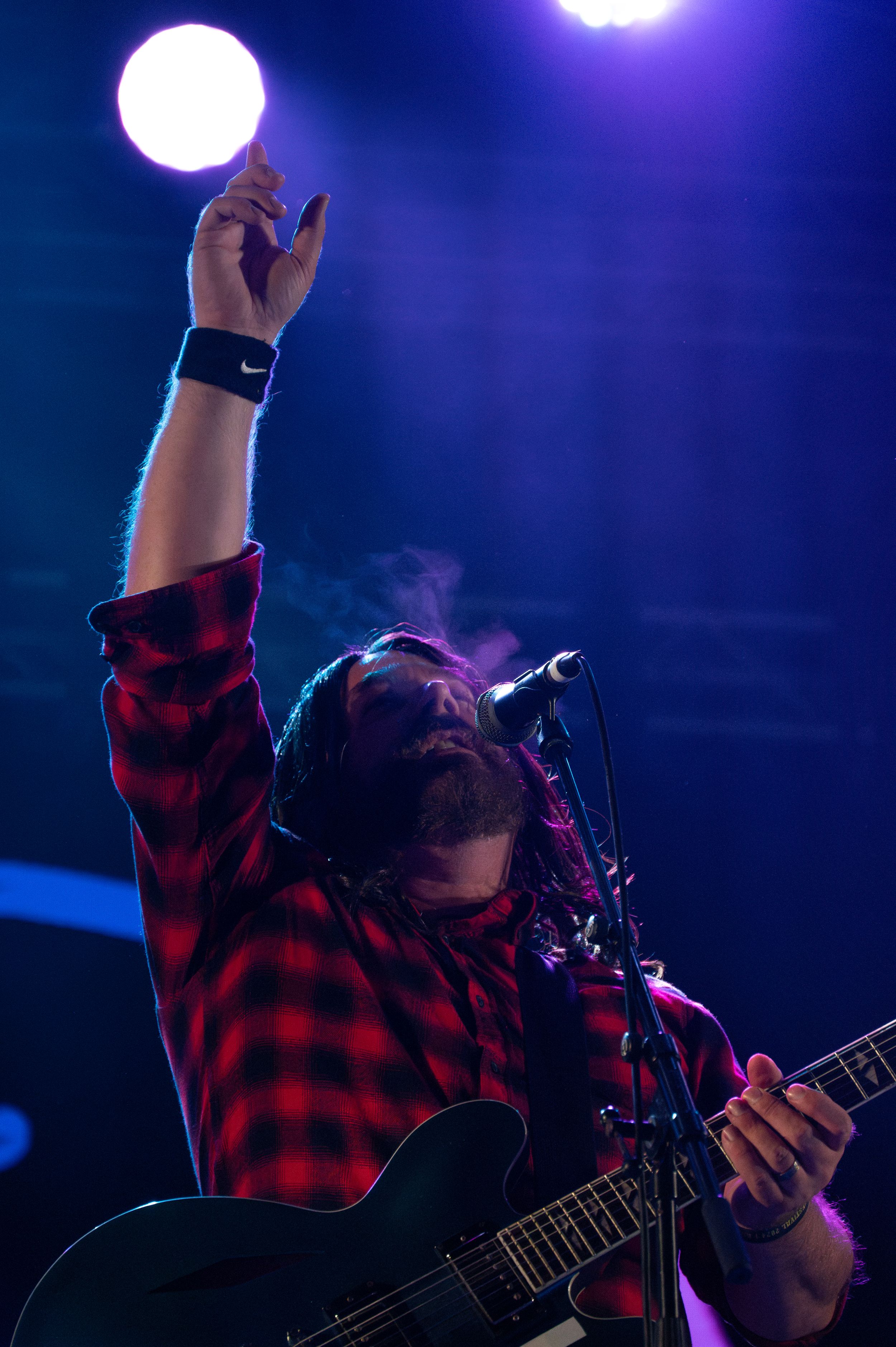 Musician in red plaid shirt raises arm during performance with dramatic blue stage lighting.