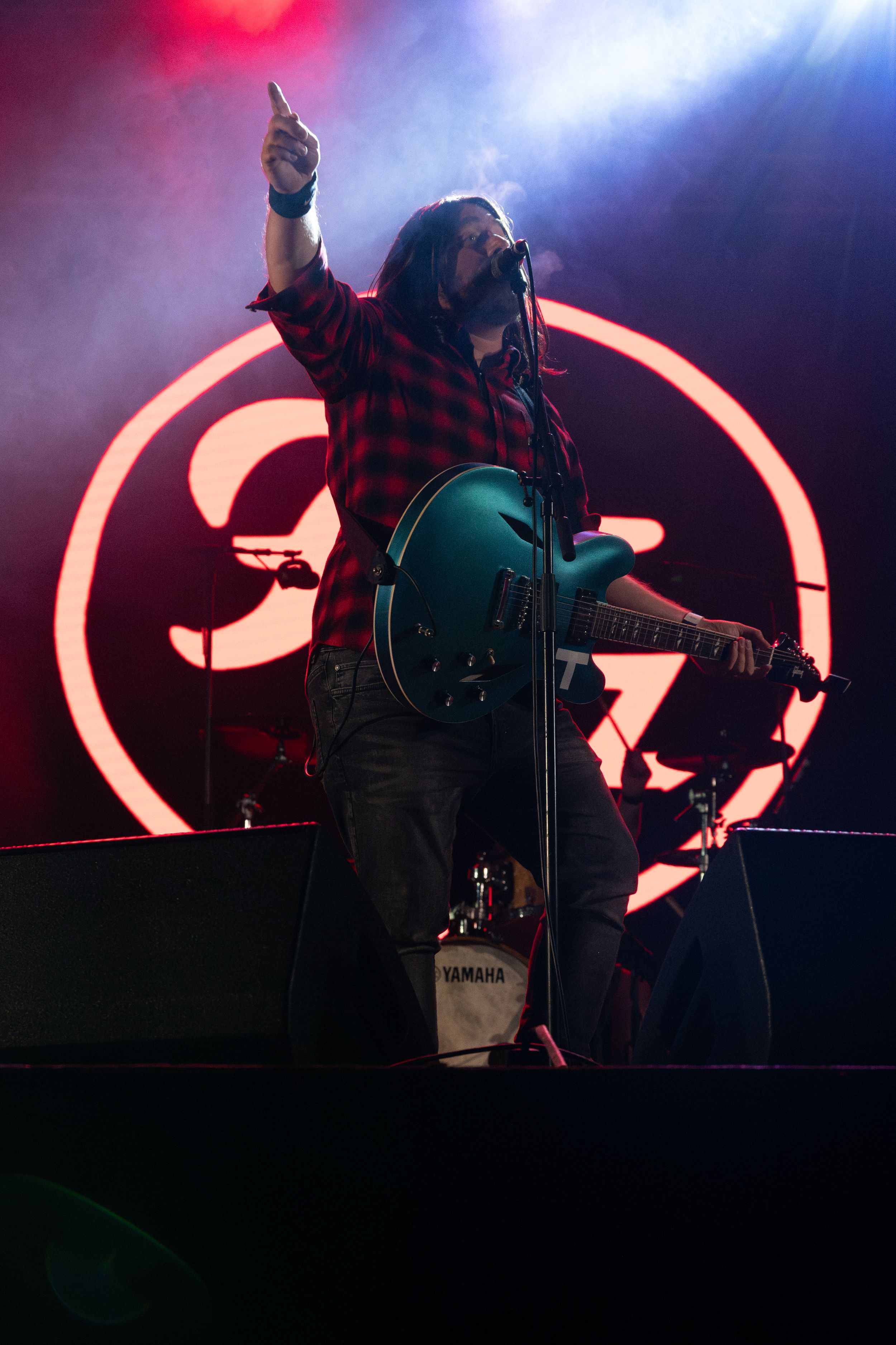 Performer in plaid shirt plays guitar in front of large illuminated circular logo backdrop.